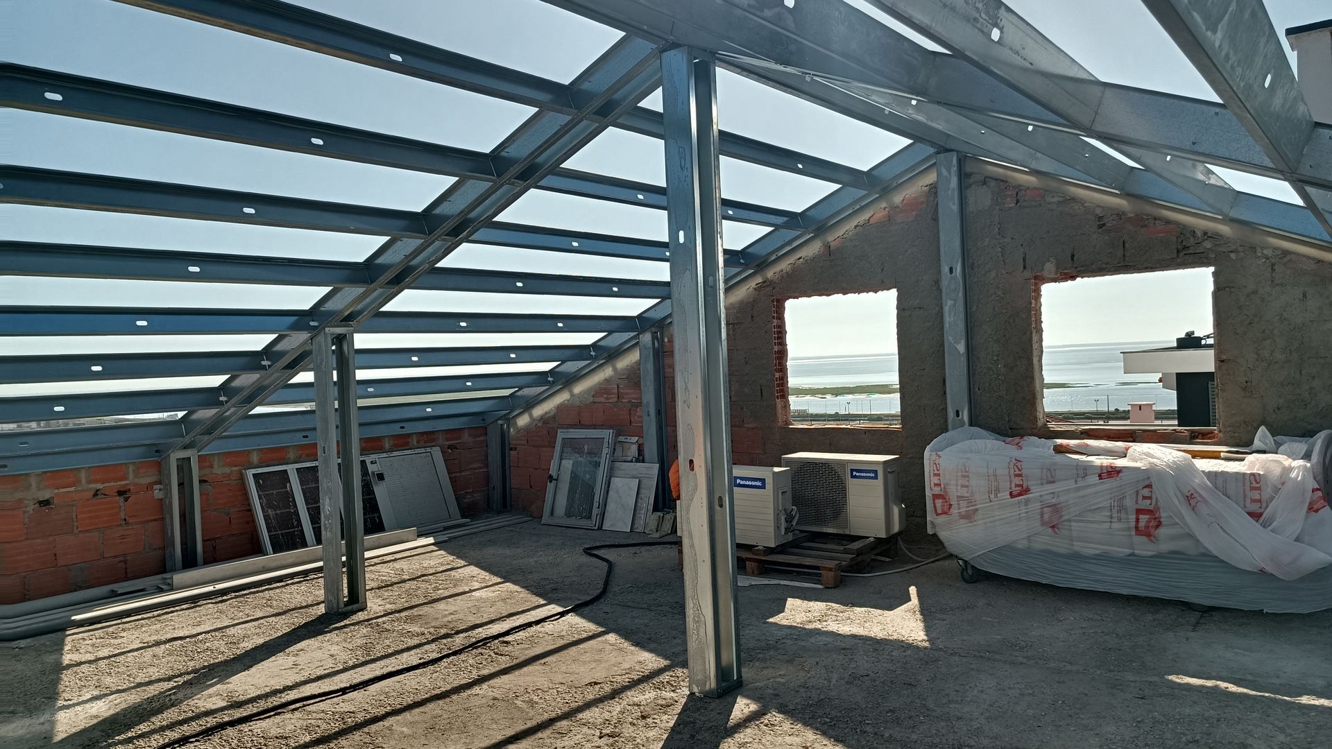 Interior de um edifício em construção, mostrando a estrutura metálica do telhado, paredes de tijolo e janelas.