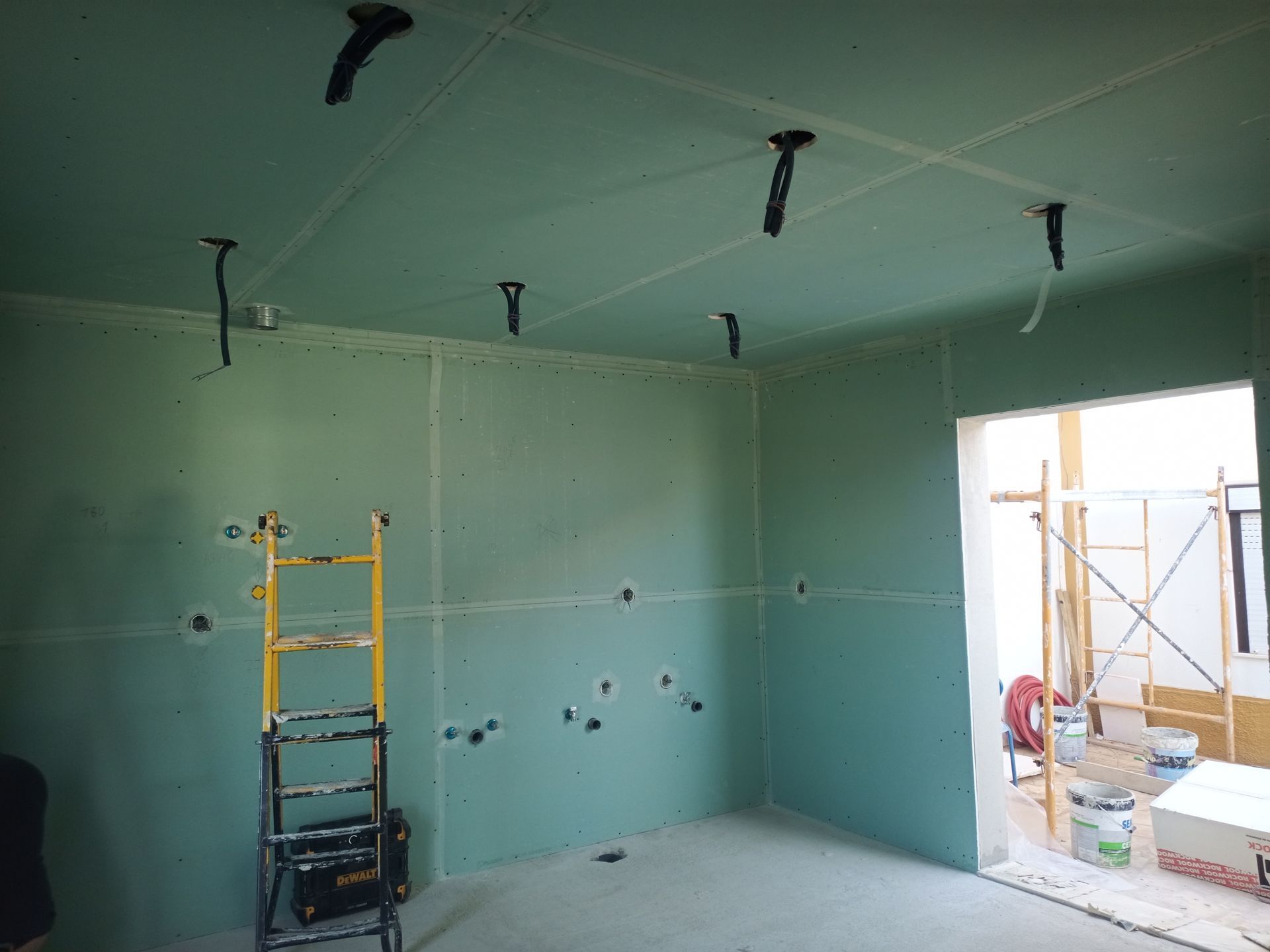 Sala em construção com drywall verde e fiação exposta, escada presente, porta aberta.