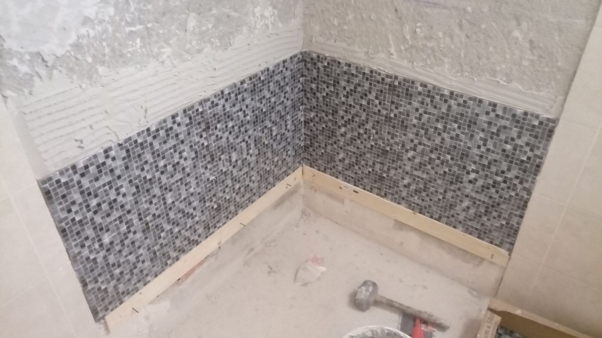 Canto do chuveiro em construção, parede revestida com azulejos de mosaico, moldura de madeira e ferramentas no chão.