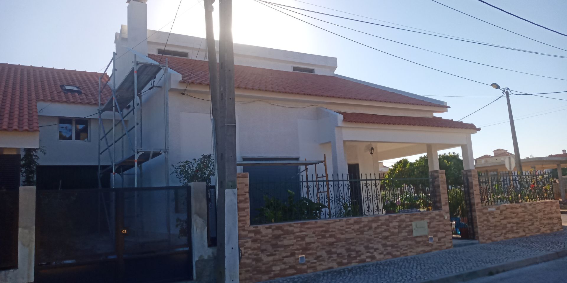 Casa branca com telhado de telha vermelha, pátio coberto, parede de tijolos e fios de energia contra um céu azul.