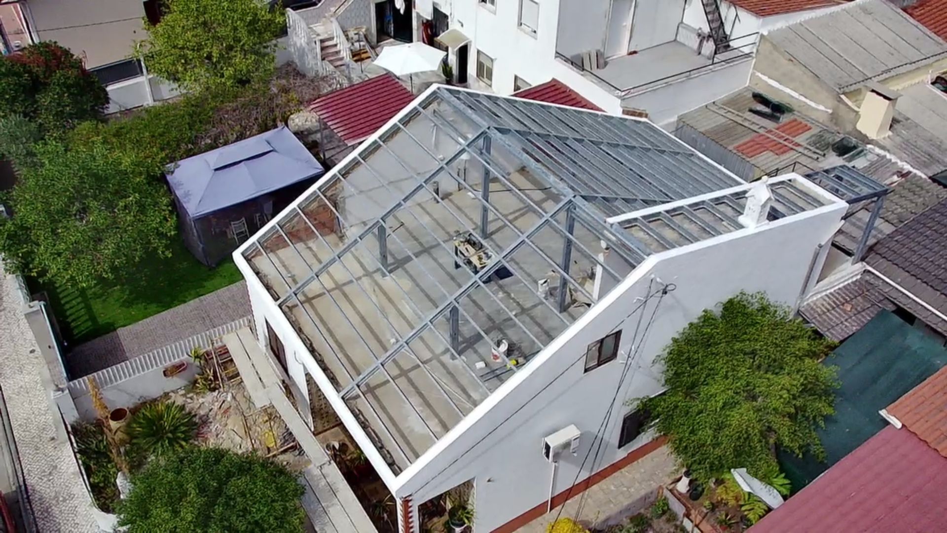 Casa branca com estrutura de telhado de vidro, rodeada por outros edifícios e vegetação.