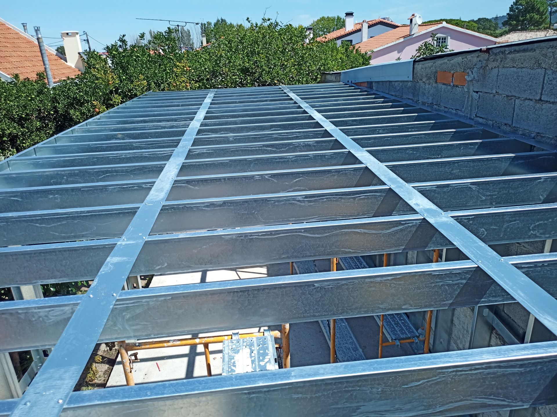 Construção com estrutura metálica para telhado, ao ar livre, sobre uma estrutura de tijolos parcialmente construída, sob forte luz solar.