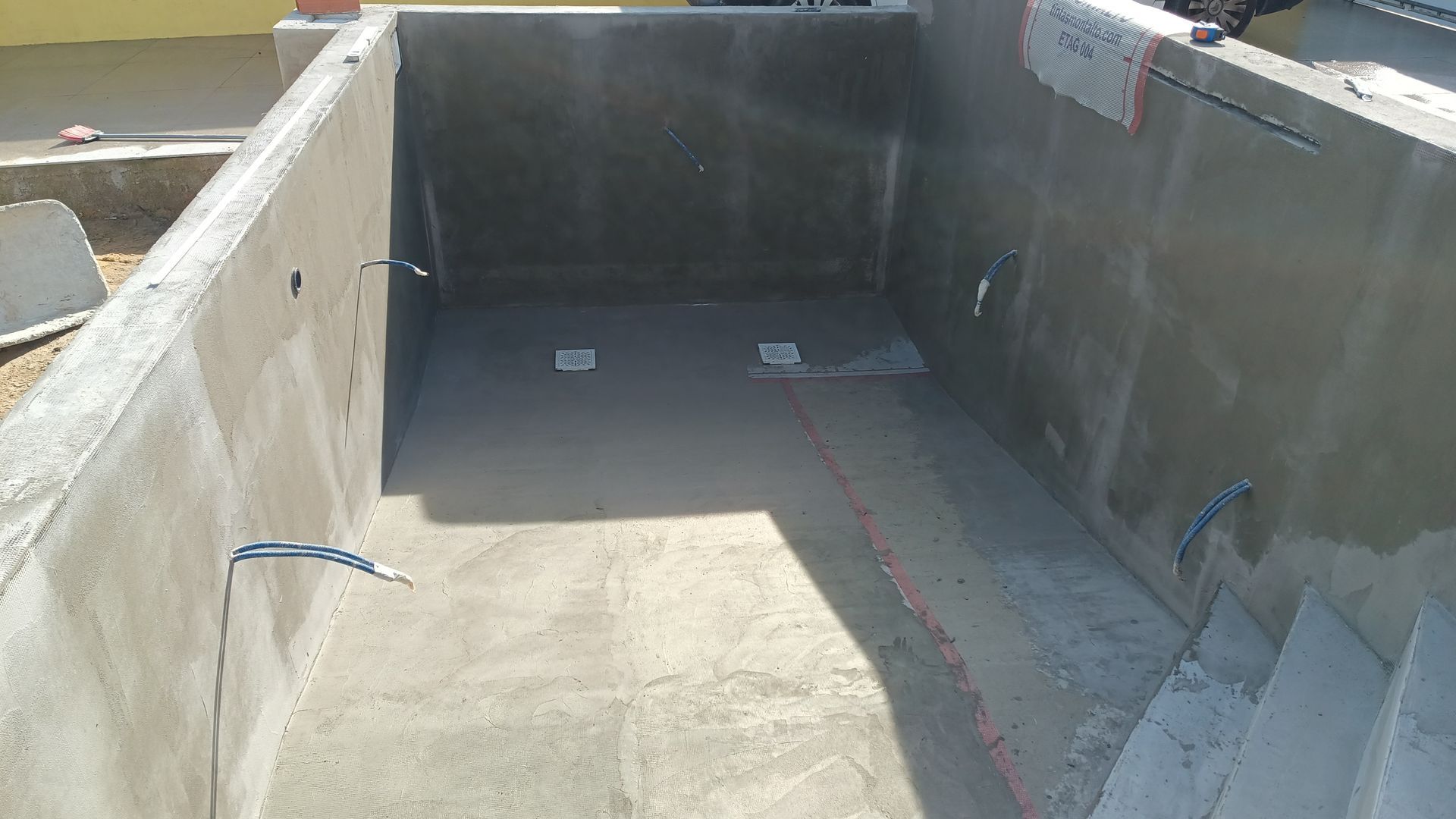 Piscina de concreto vazia em construção; paredes e piso cinza, encanamento visível.