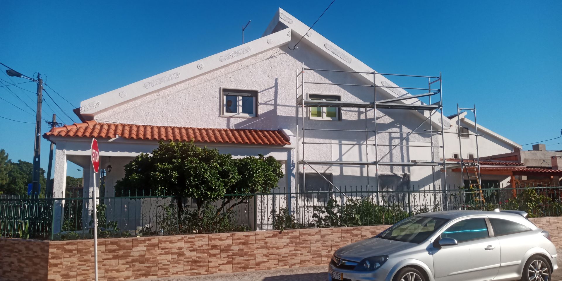 Casa branca com telhado de telhas laranja e andaimes, carro estacionado em frente em um dia ensolarado.