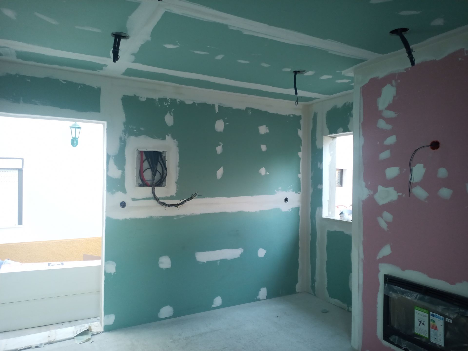 Interior de um cômodo em construção com paredes de gesso acartonado nas cores verde e rosa. Fiação elétrica exposta.