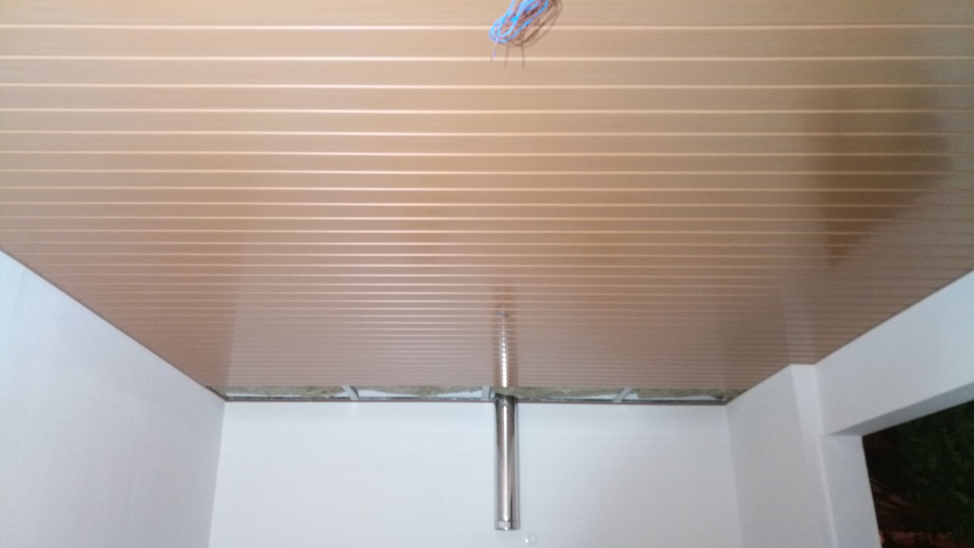 Teto com painéis bege acima de paredes brancas com um cano prateado estendendo-se para baixo.