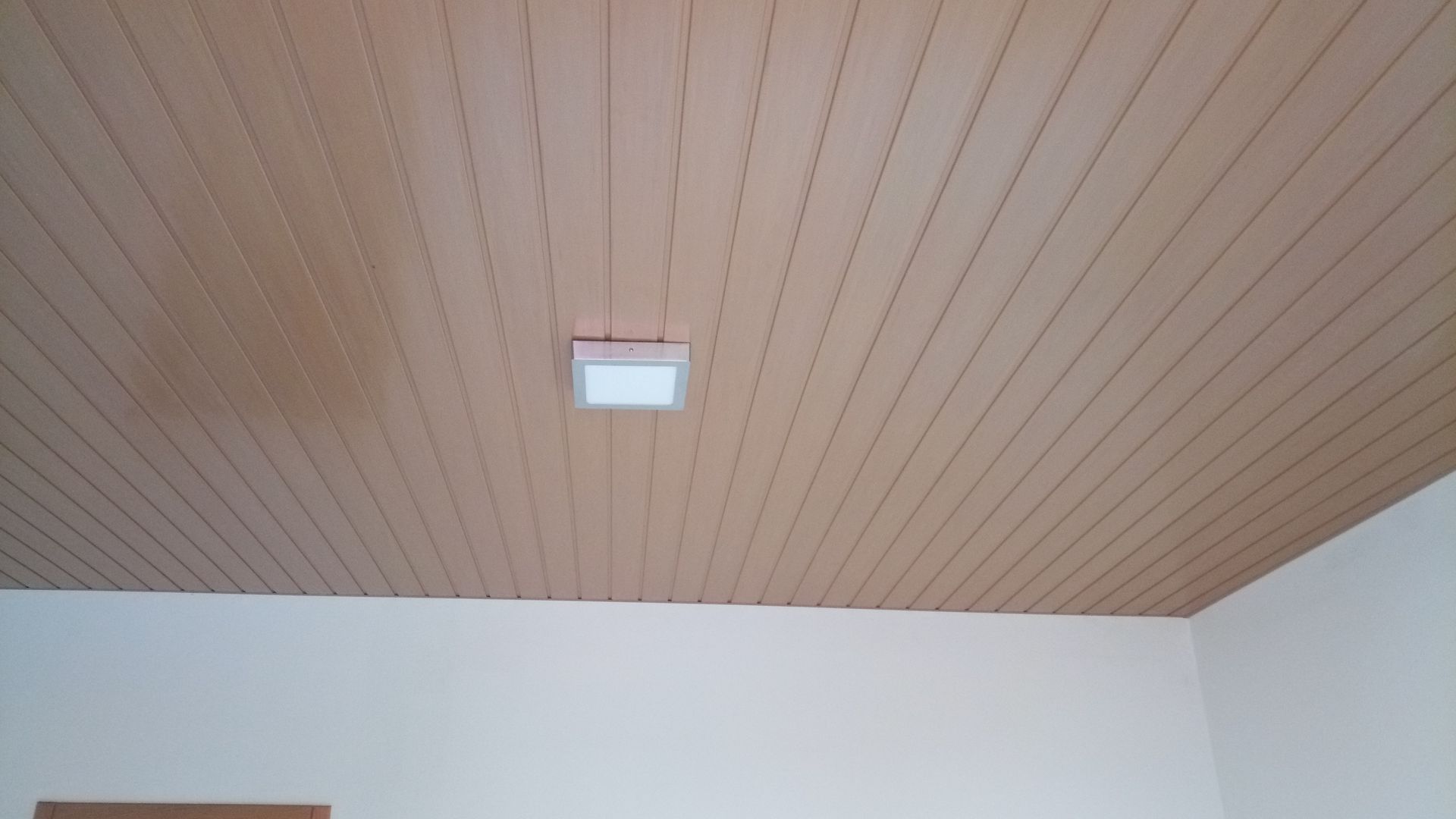 Teto com painéis de madeira e luminária quadrada embutida. Paredes brancas na parte inferior.