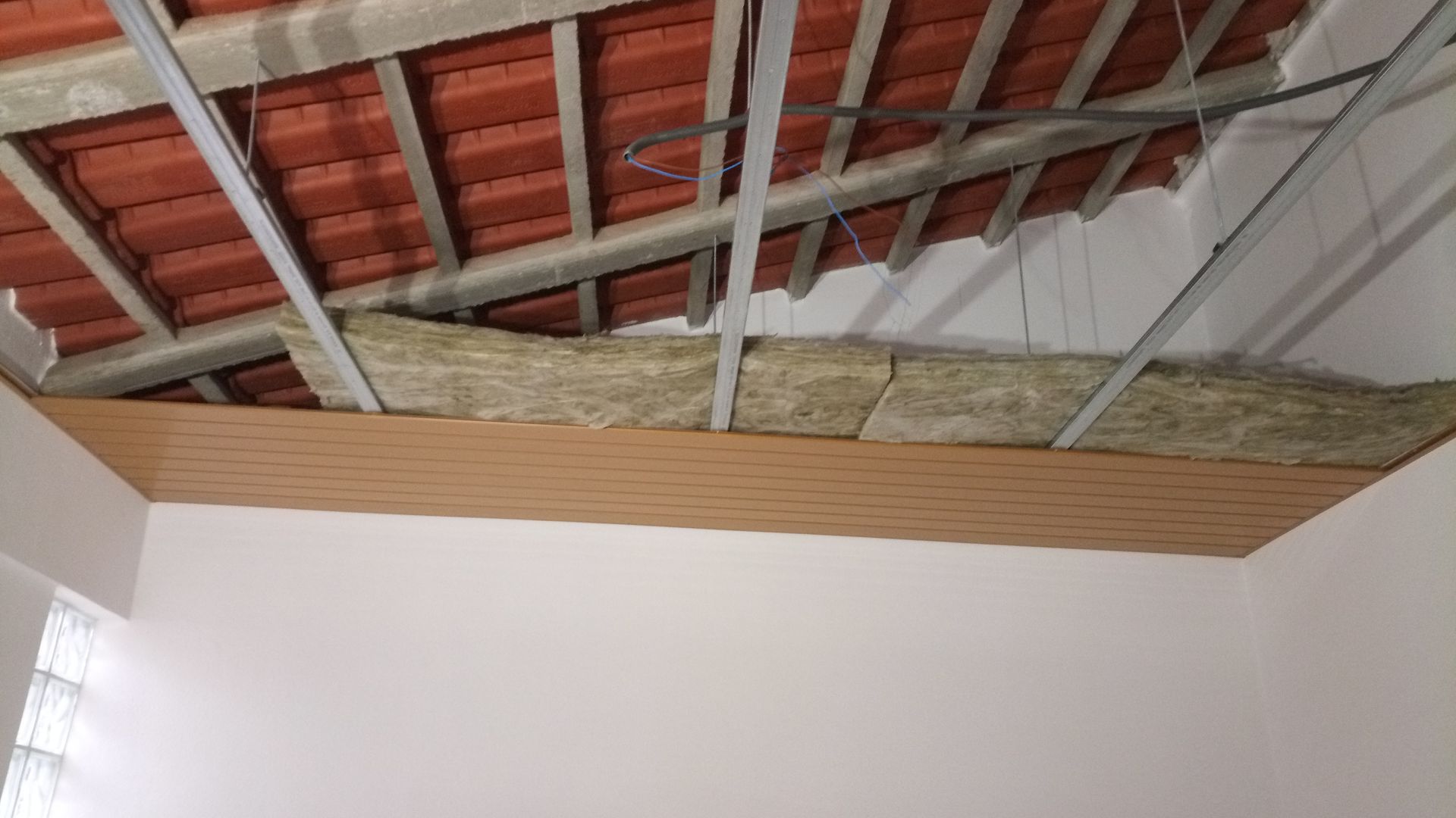 Construção do teto interior: cobertura de telha aparente, estrutura metálica, isolamento e painéis de madeira sobre uma parede branca.