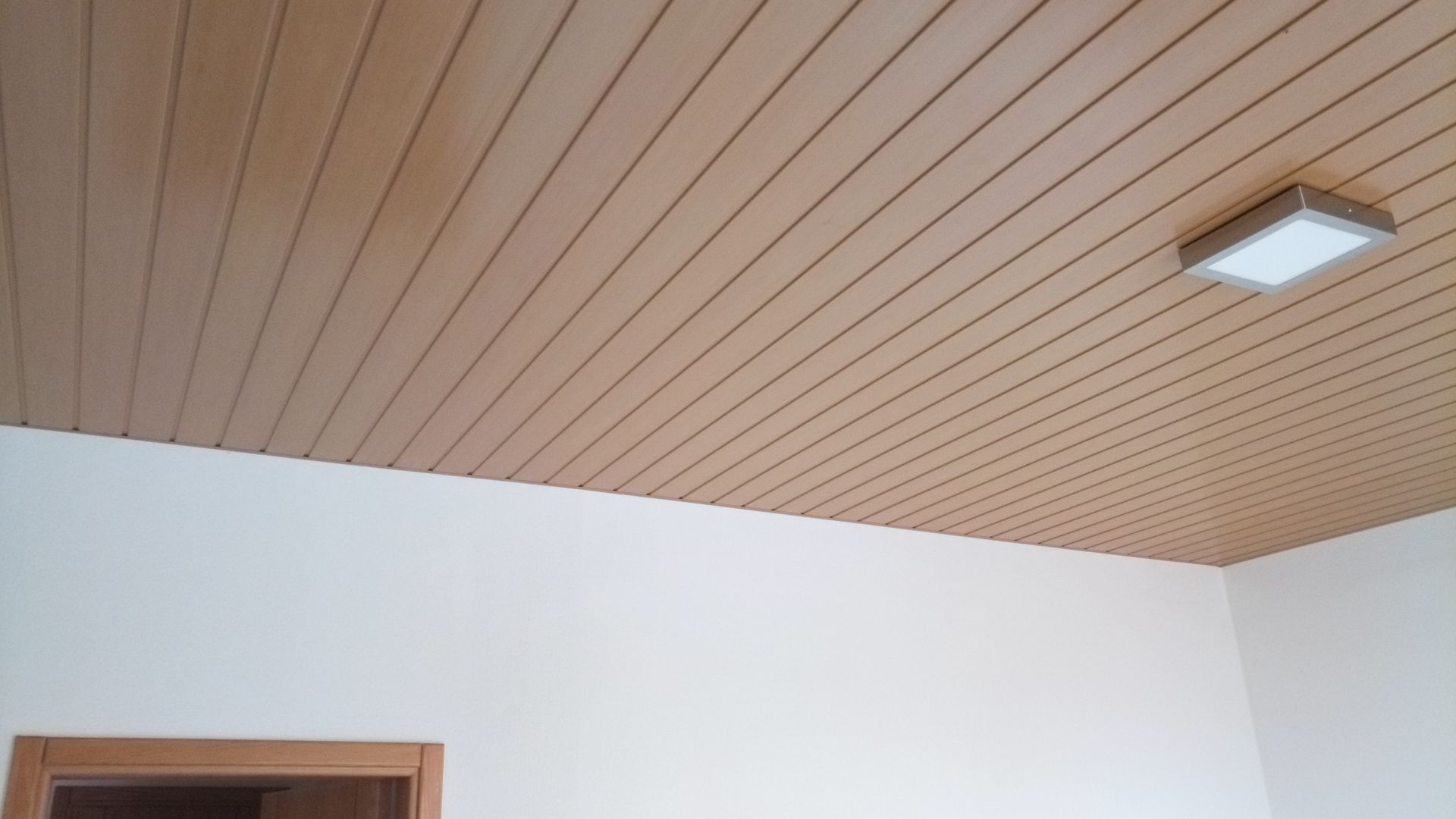 Teto com painéis de madeira, luminária retangular, canto com paredes brancas e porta.