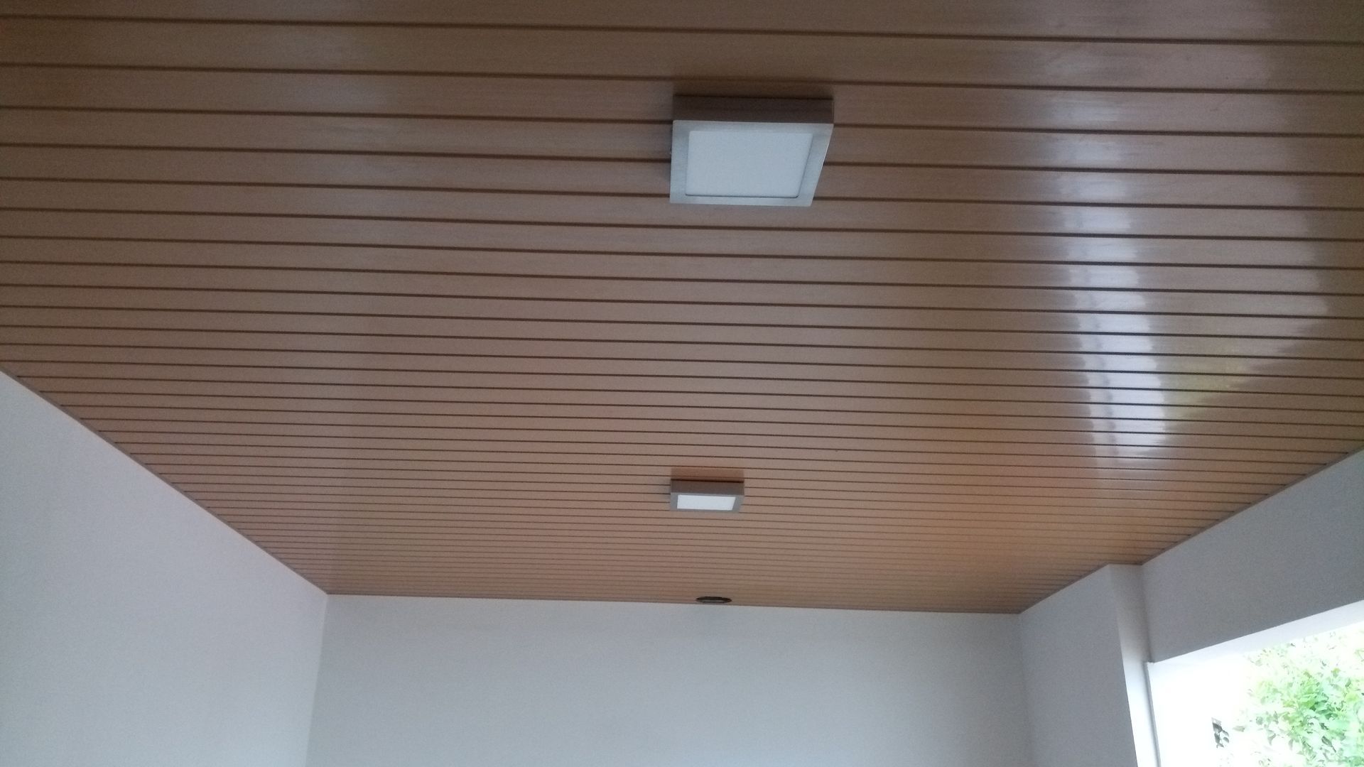 Teto ripado de madeira com duas luminárias quadradas brancas, acima de paredes brancas.