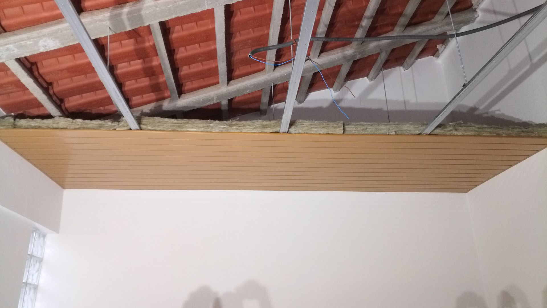 Teto com painéis de madeira marrom, sustentado por estrutura metálica, abaixo de telhado aparente de telhas vermelhas com isolamento.