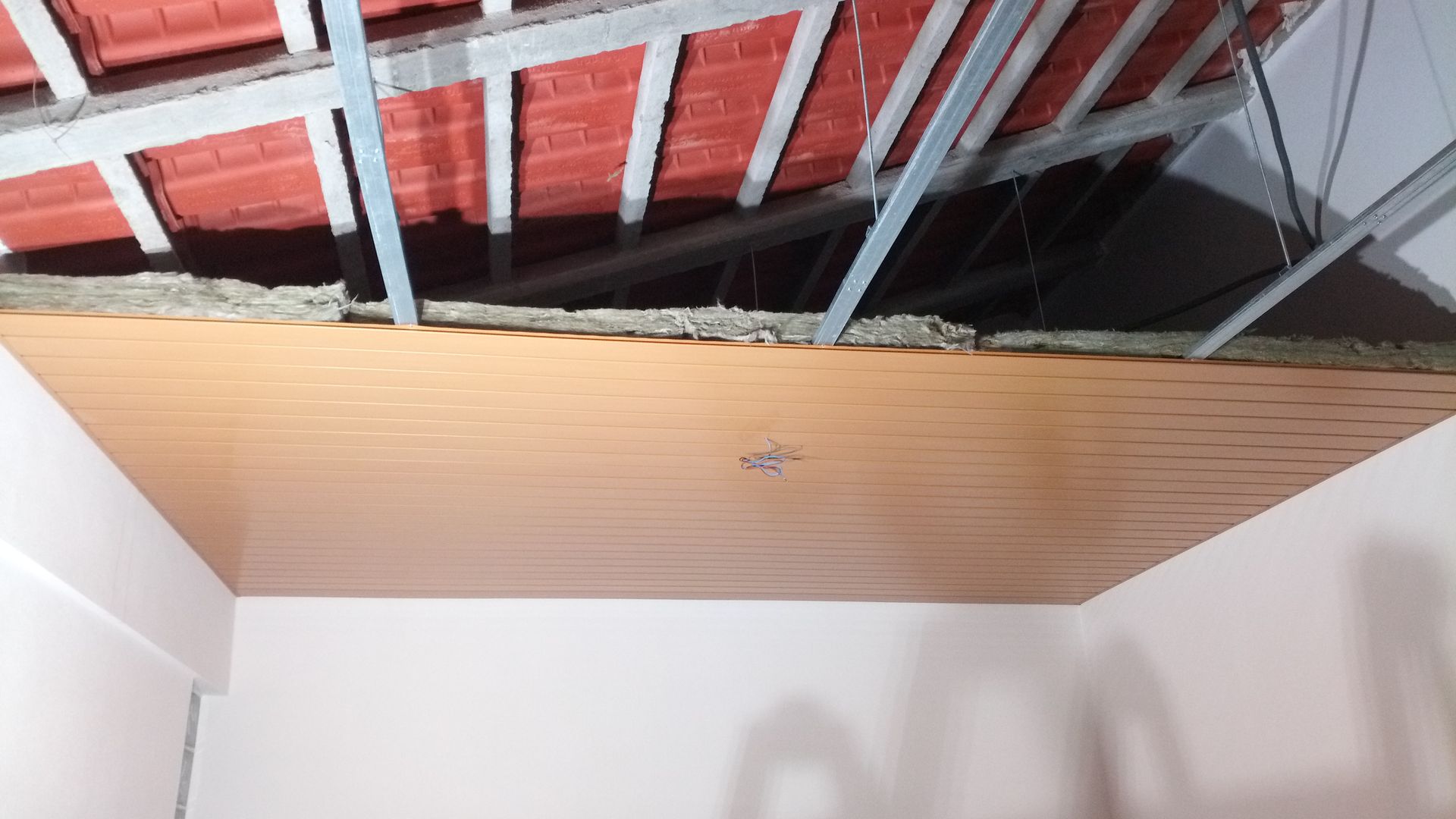Teto marrom com iluminação embutida, isolamento e estrutura metálica que sustenta telhas vermelhas.