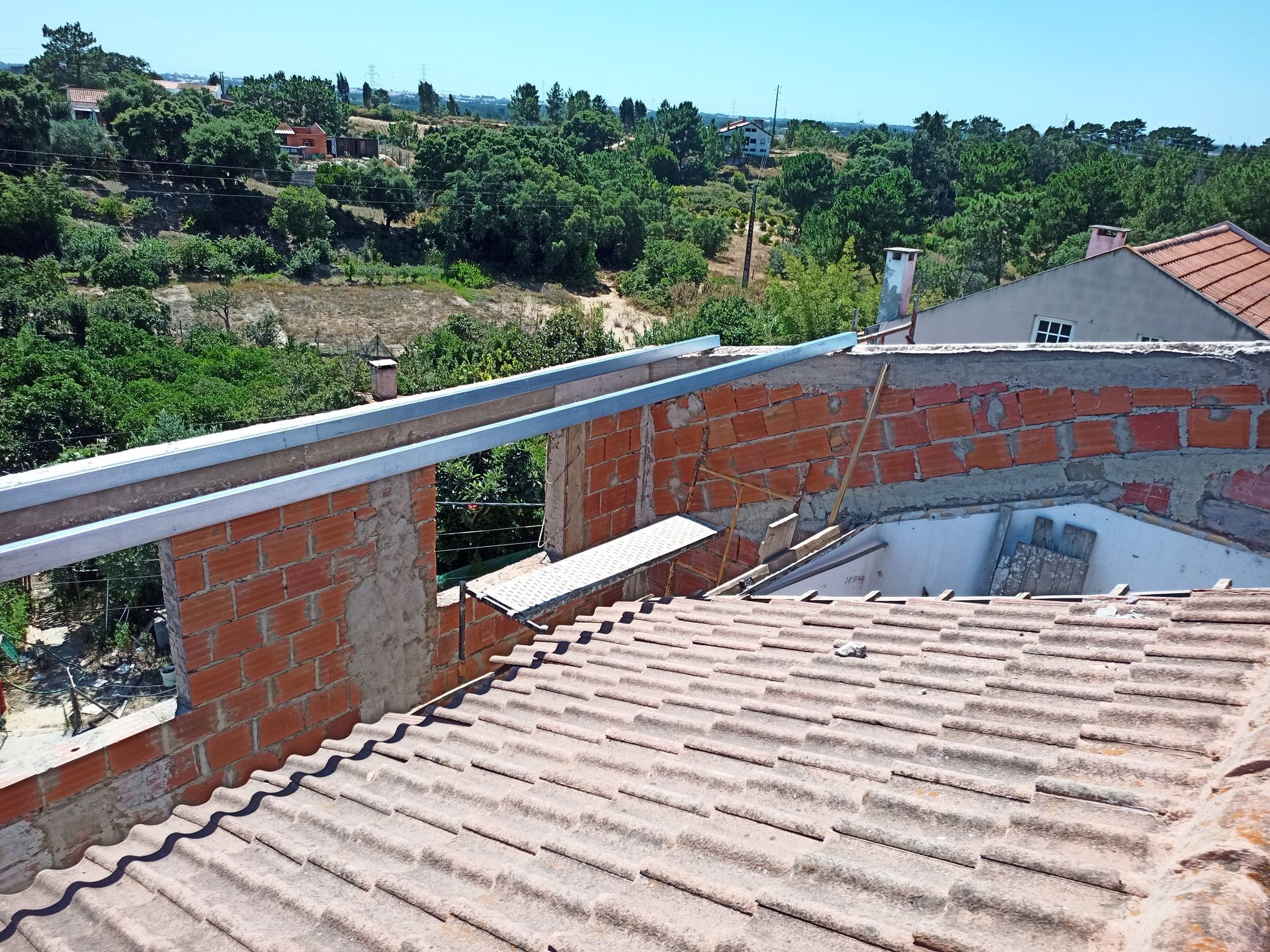 Terraço com parede de tijolos vermelhos, calha de metal e telhado de telha cerâmica, tendo como pano de fundo árvores e um céu azul límpido.
