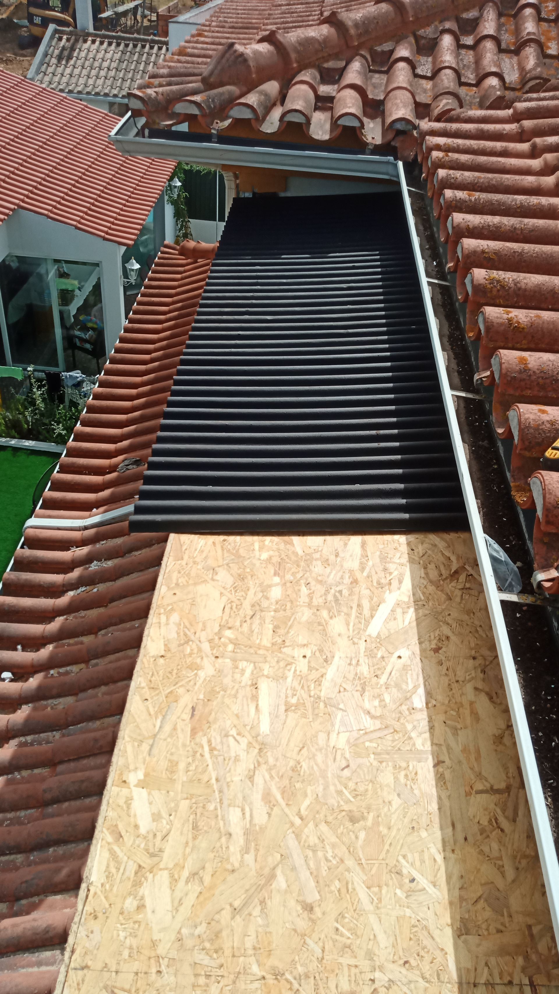 Reparo do telhado em andamento: compensado e subcobertura preta substituem as telhas. Telhado marrom e vermelho em um ambiente externo ensolarado.