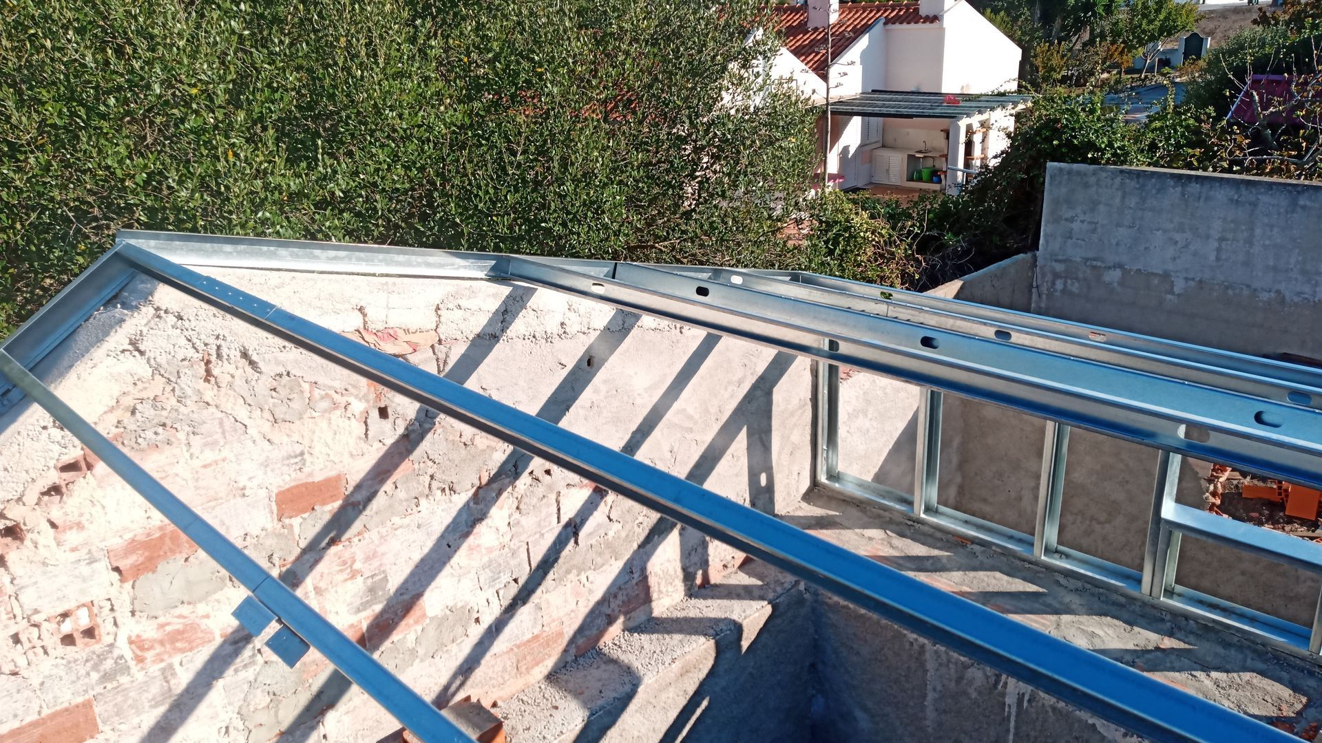 Estrutura metálica instalada sobre um telhado de tijolos. A luz do sol projeta sombras. Prédios e vegetação ao fundo.
