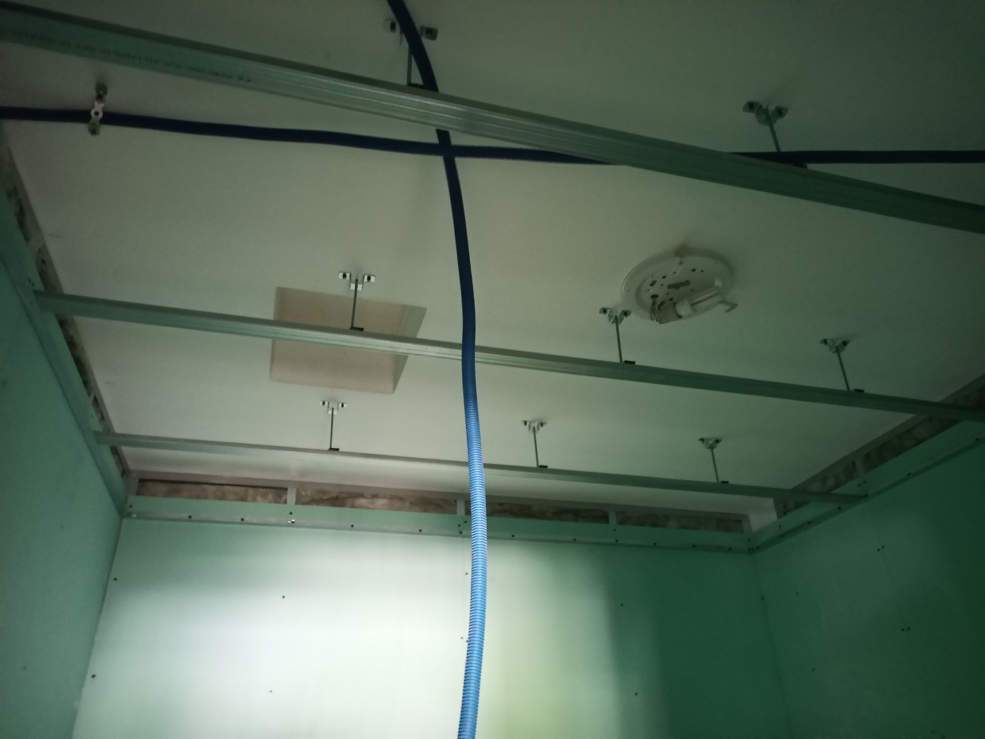 Vista interna de um cômodo em reforma. Vigas de sustentação do teto expostas, cabo azul e um trecho de drywall faltando.