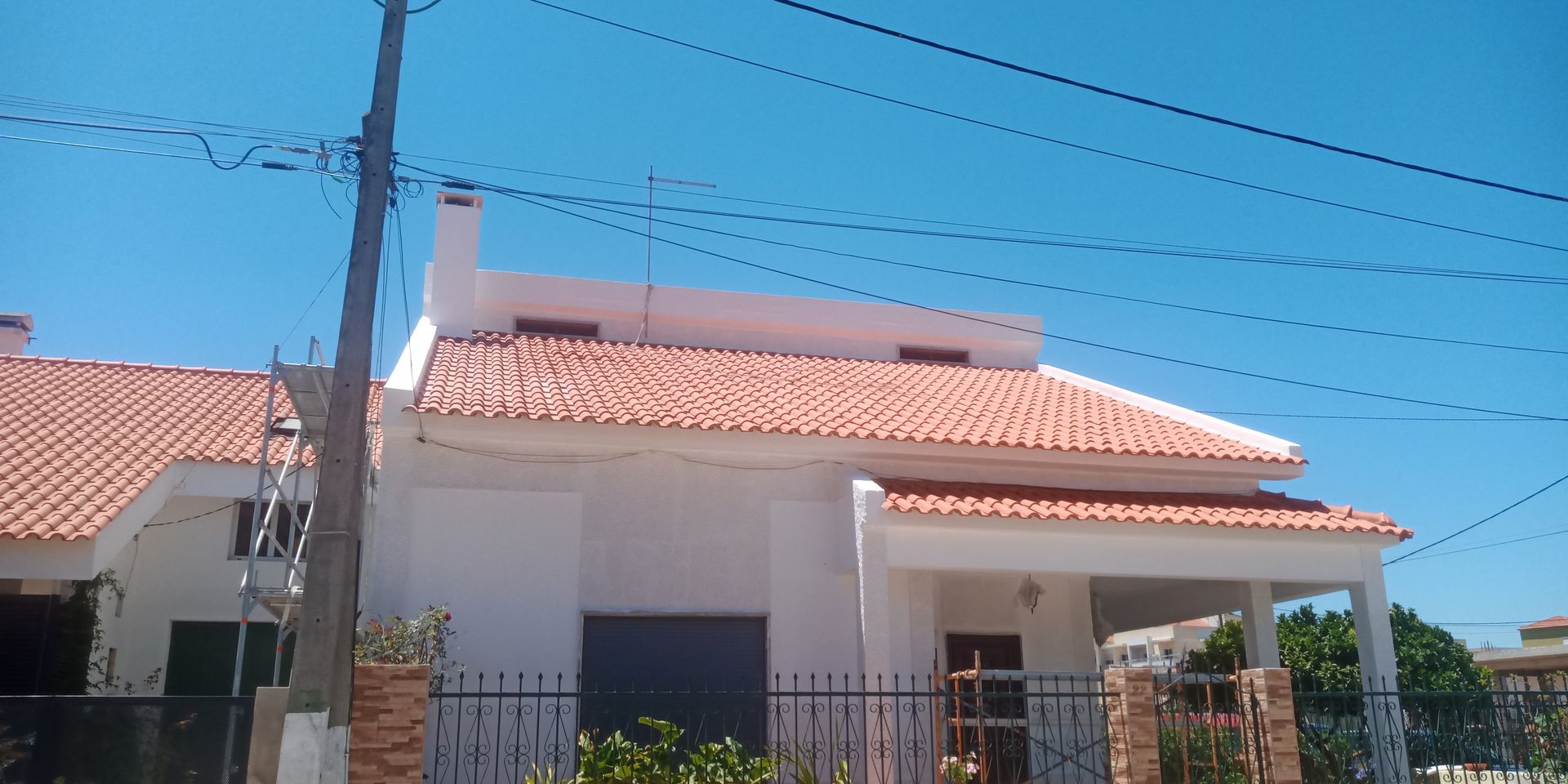 Casa branca com telhado de telha laranja, varanda, cerca e fios de energia contra um céu azul.