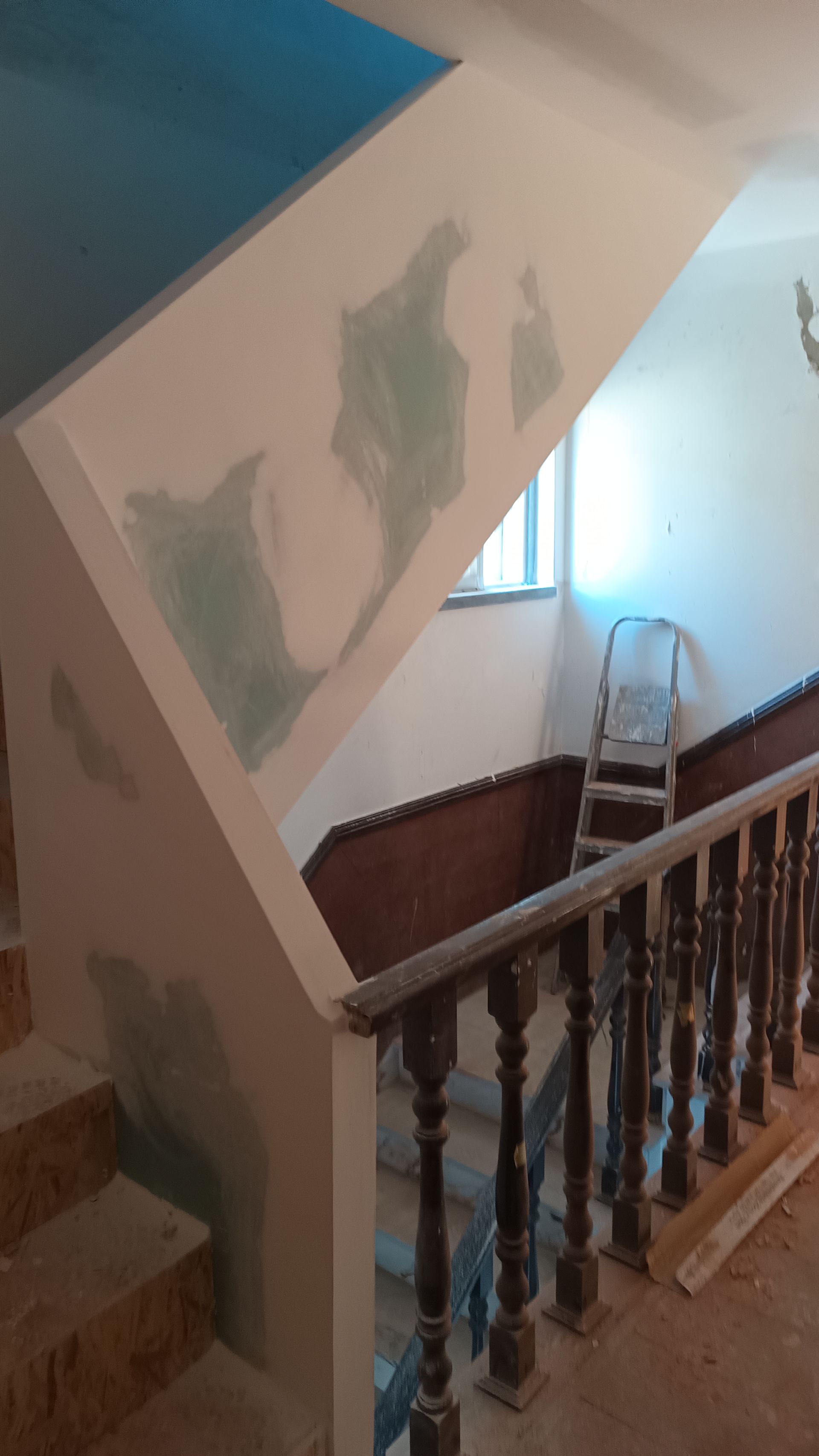 Escada com reparos na parede, corrimão e uma escada de mão perto de uma janela. Tons de branco e marrom predominam.