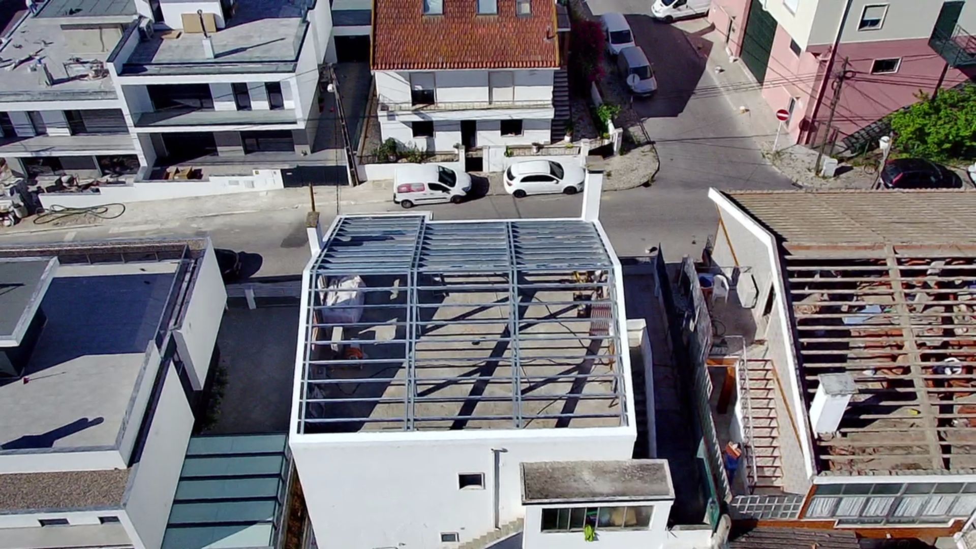 Vista aérea de um edifício branco em construção com estrutura metálica exposta no telhado, cercado por outros edifícios e uma rua.