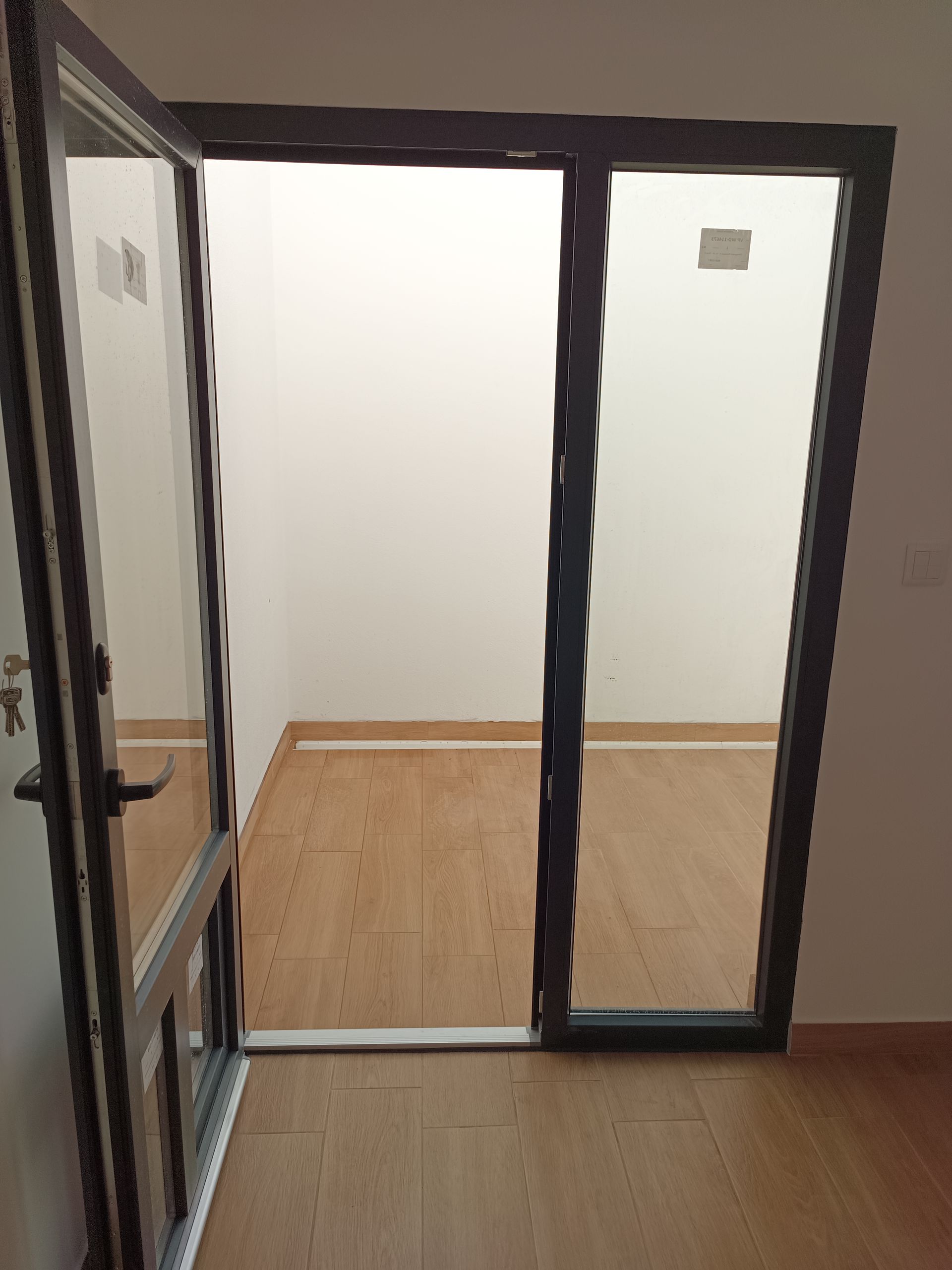 Porta de vidro com moldura escura, parcialmente aberta, que dá acesso a uma sala com piso de madeira e paredes brancas.