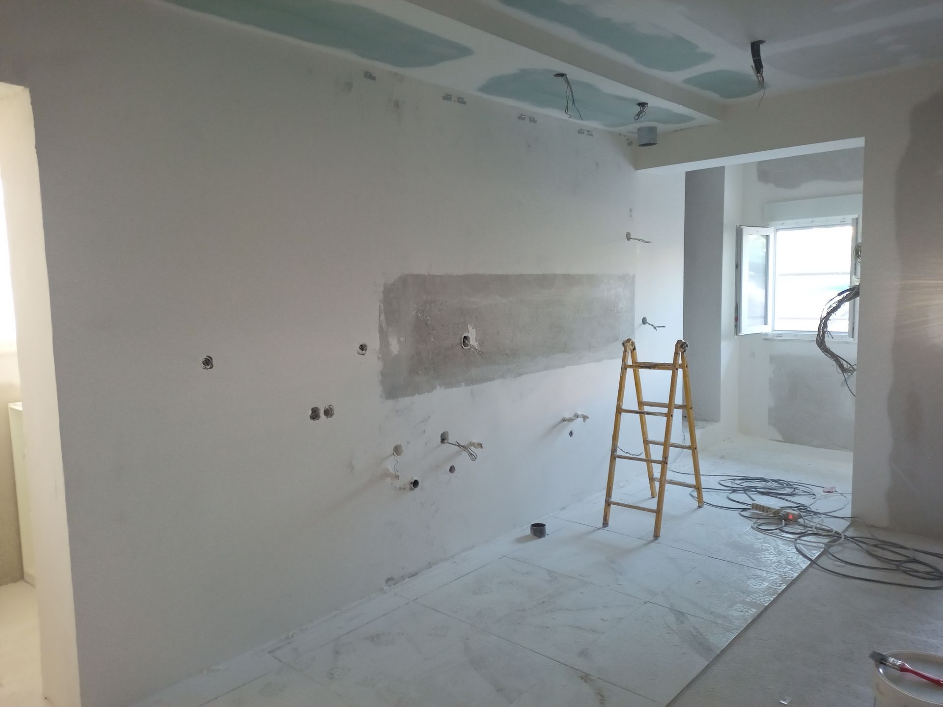 Interior de um cômodo em construção, com paredes de gesso e fiação exposta. Uma escada está próxima.