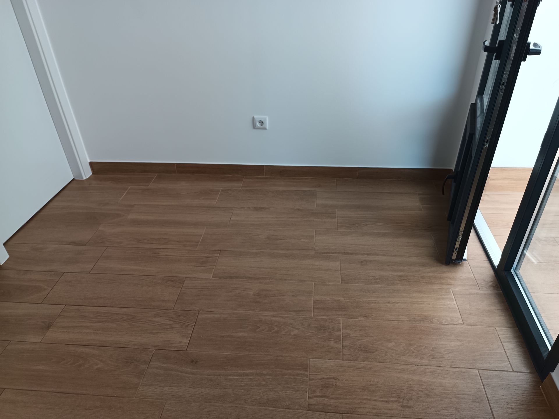 Quarto vazio com piso imitando madeira, paredes brancas, porta à esquerda e porta de vidro deslizante à direita.