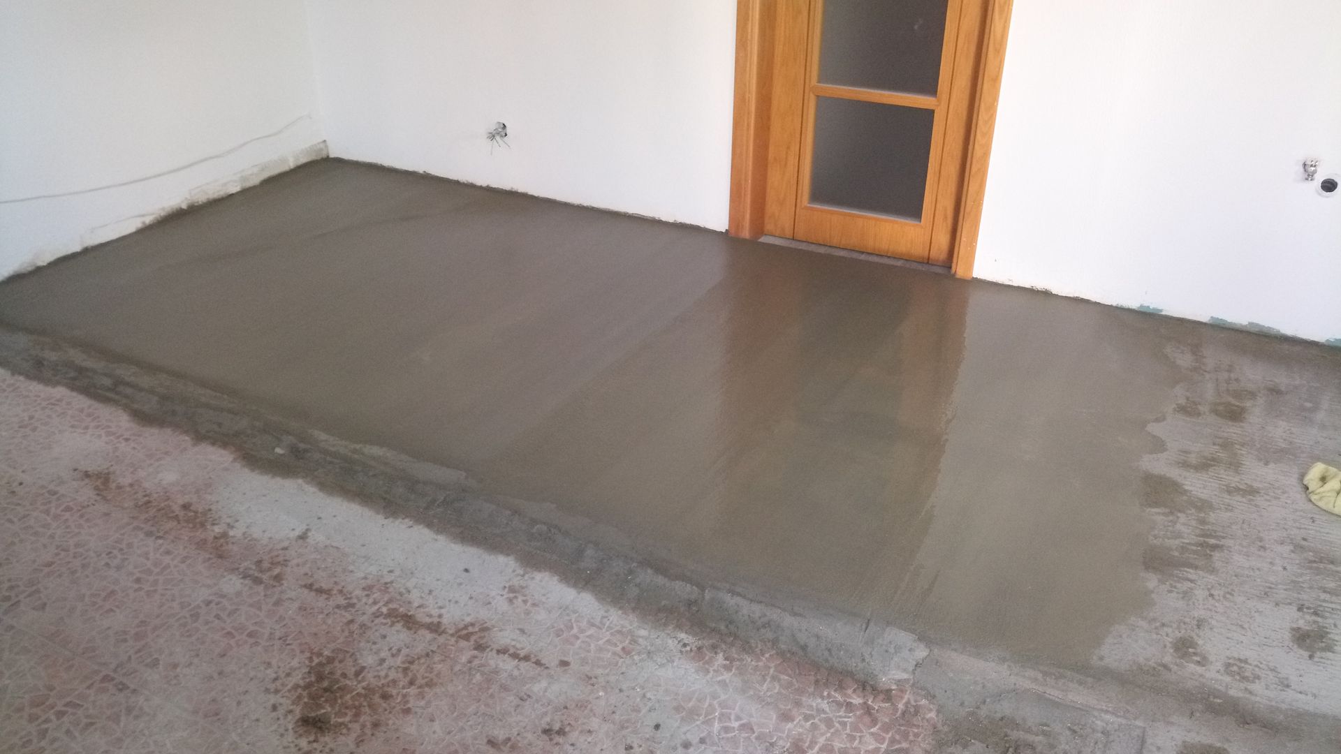 Piso de concreto recém-moldado em um espaço interior, refletindo a luz, ao lado de uma porta e paredes em tom branco-sujo.