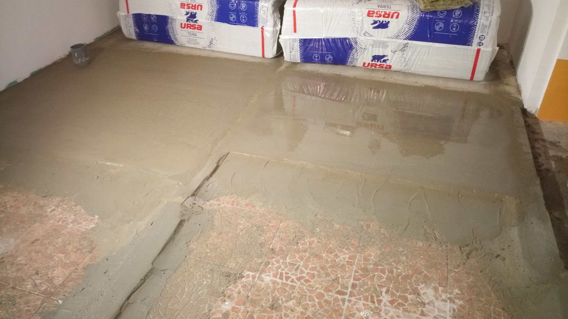 Piso de concreto sendo despejado, algumas seções já secas. Dois sacos de material de construção ao fundo.