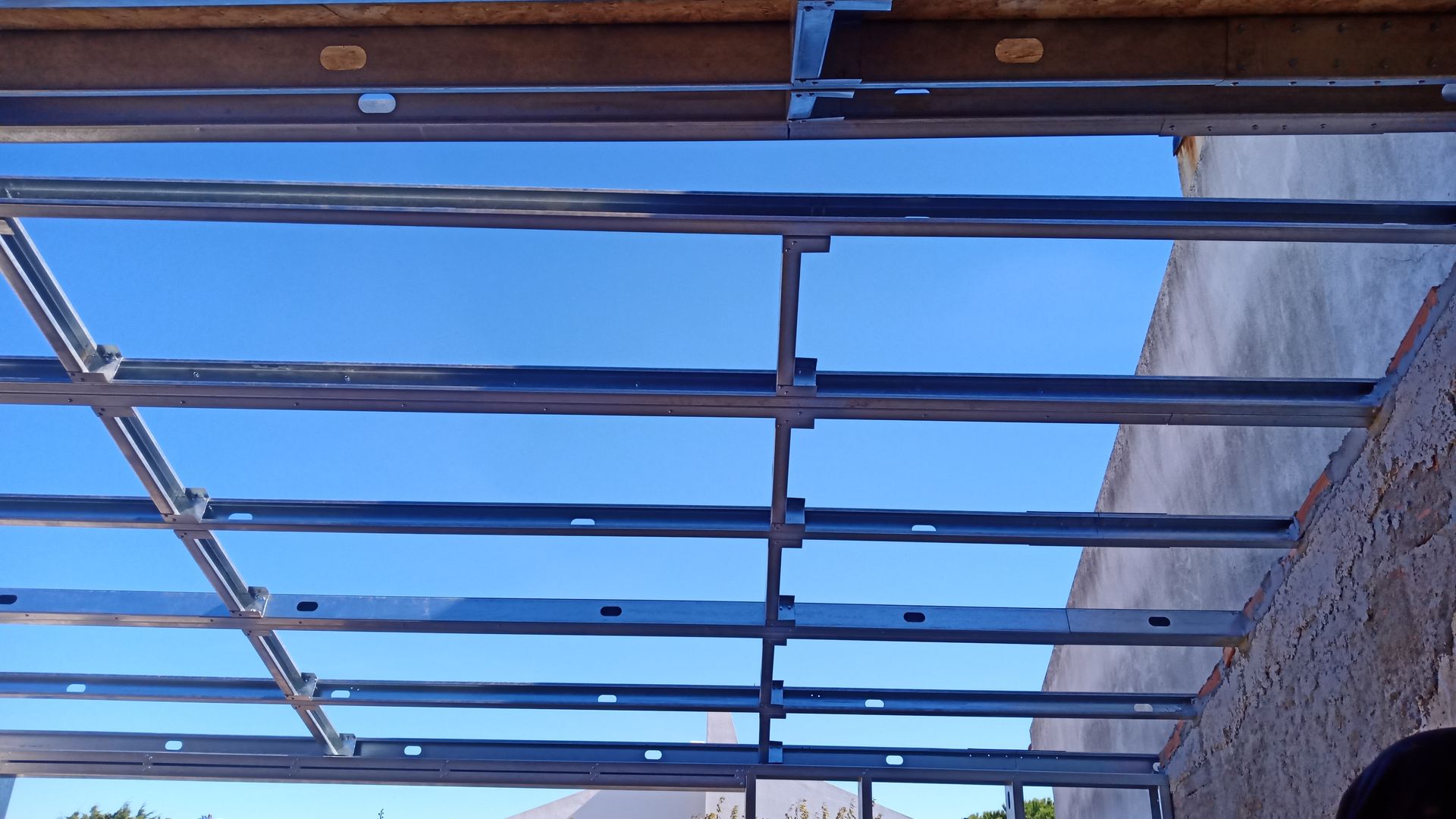 Estrutura metálica para um telhado sob um céu azul, parcialmente construída sobre uma parede.