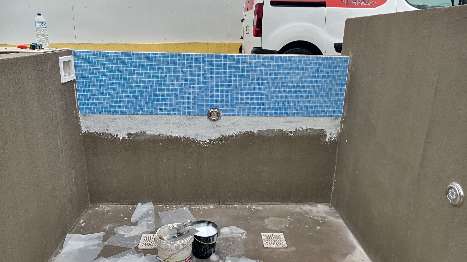 Piscina em construção, com azulejos azuis, paredes de concreto cinza e encanamento à mostra.