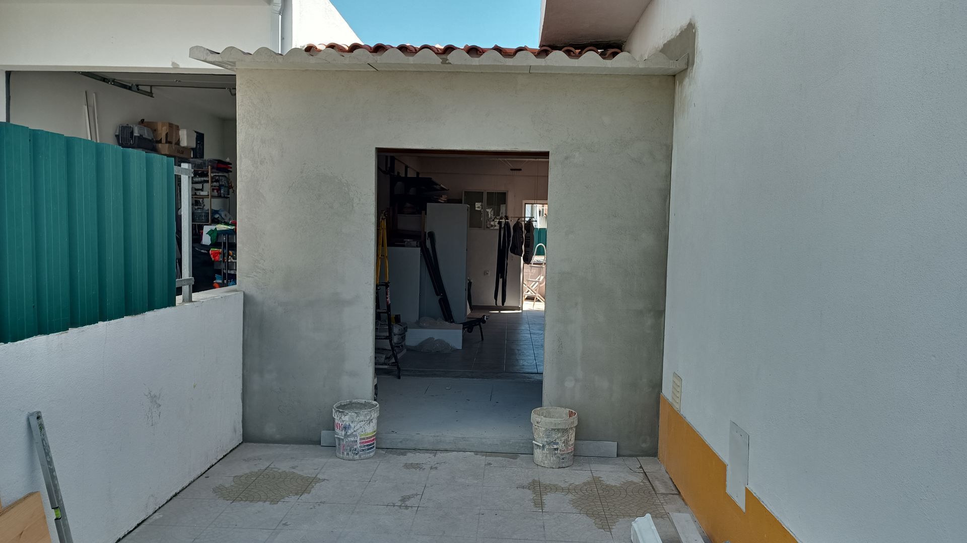 Entrada aberta de um prédio cinza e inacabado. Parede branca à direita. Cerca verde à esquerda.