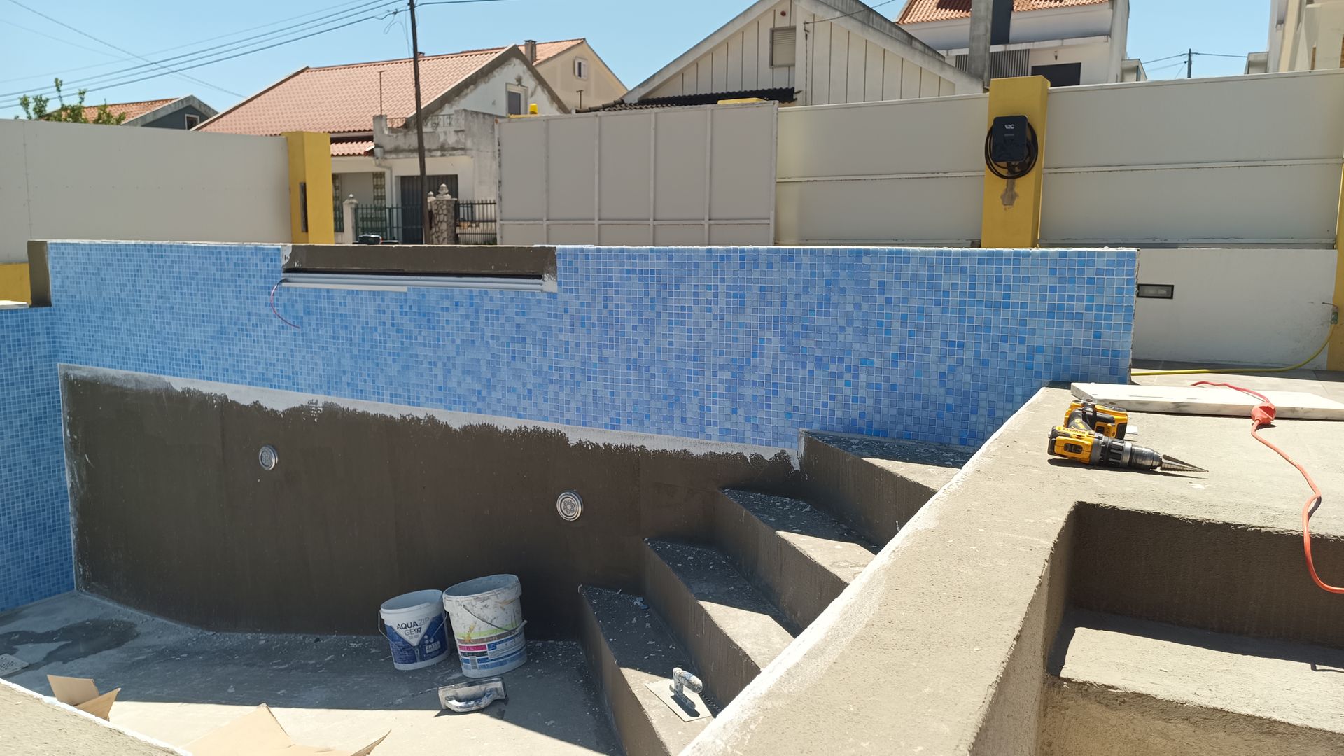 Piscina em construção, com azulejos azuis sendo instalados nas paredes. Degraus, baldes e ferramentas também são visíveis.
