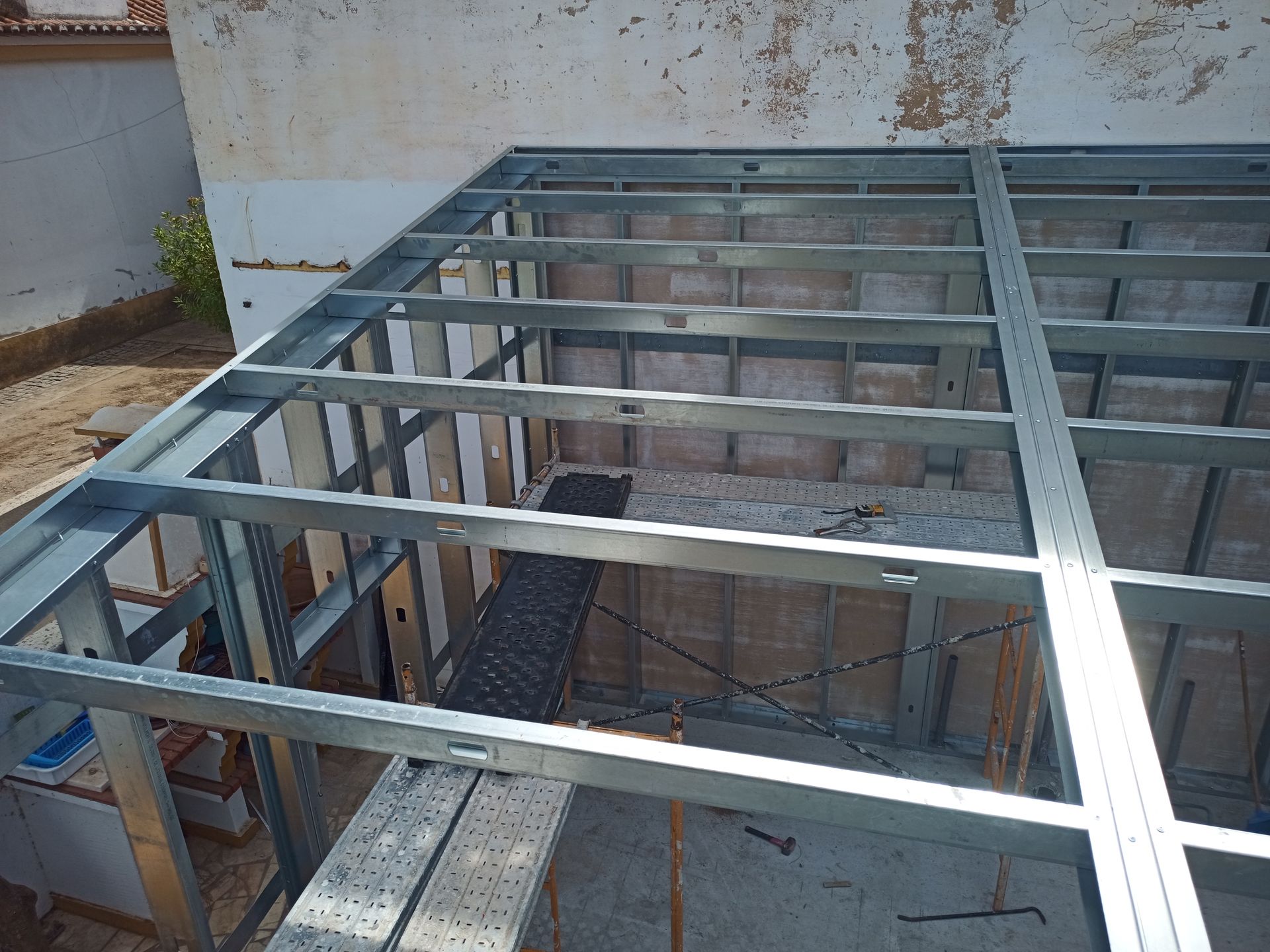Estrutura metálica de um telhado em construção contra uma parede bege desgastada.