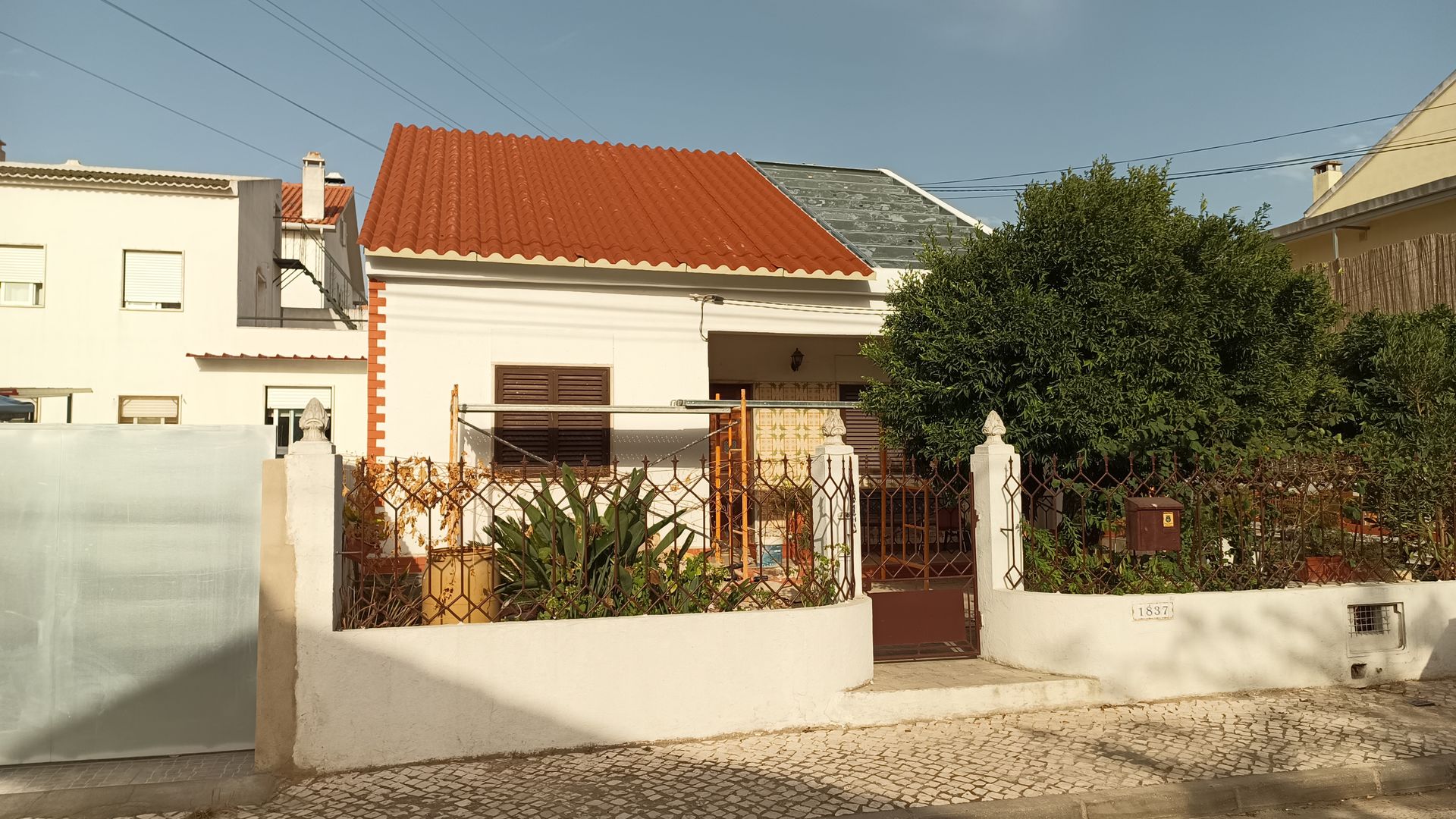 Pequena casa branca com telhado laranja, cercada por um muro baixo e um portão de ferro forjado, com arbustos na frente.