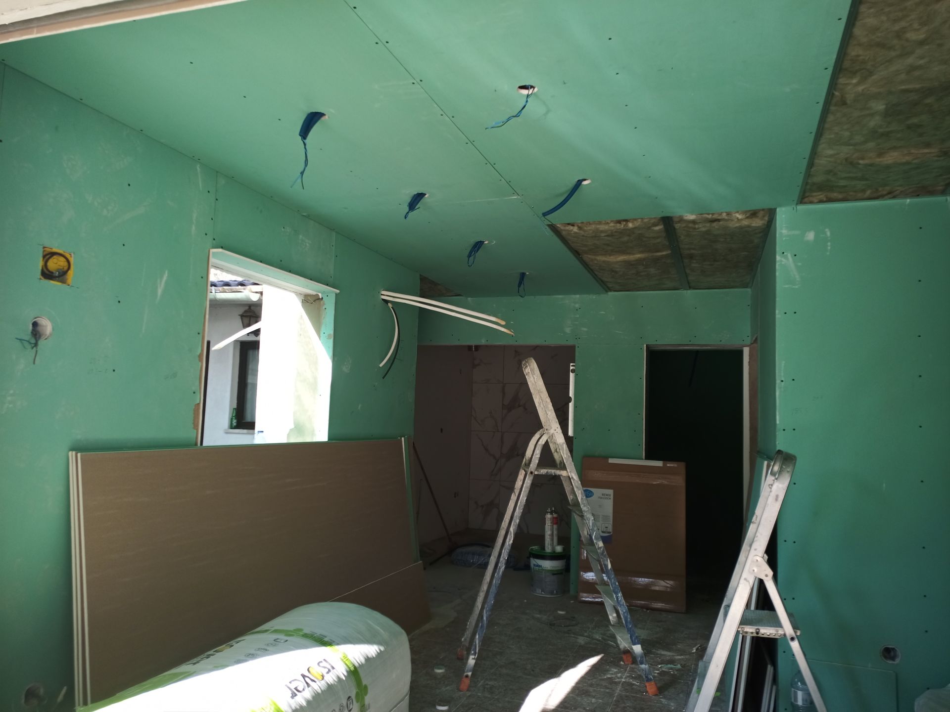 Interior de uma sala em construção, com paredes de gesso verde, fios expostos e escadas.