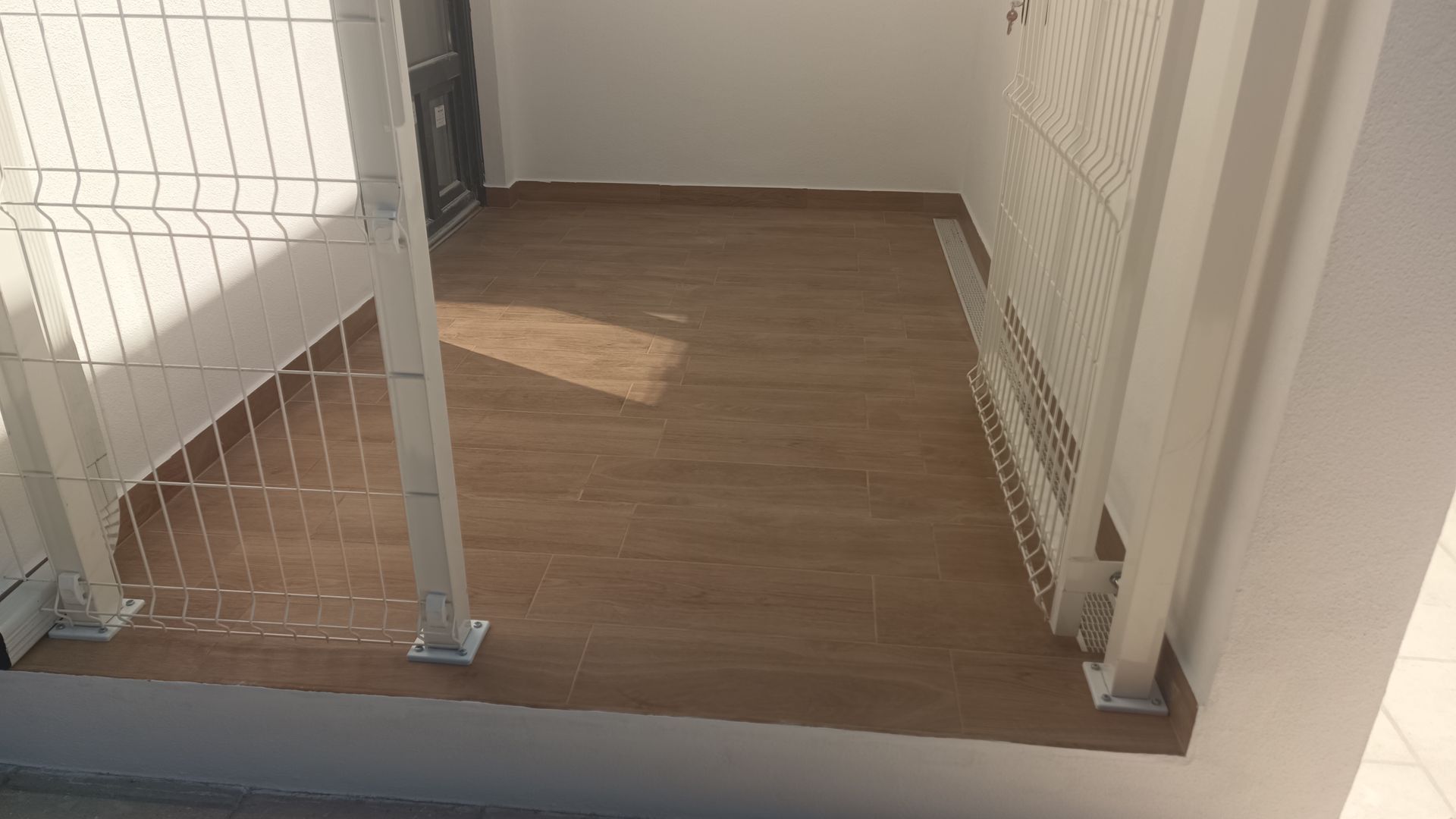 Pequena varanda com piso imitando madeira e guarda-corpo de metal branco. Iluminação abundante.