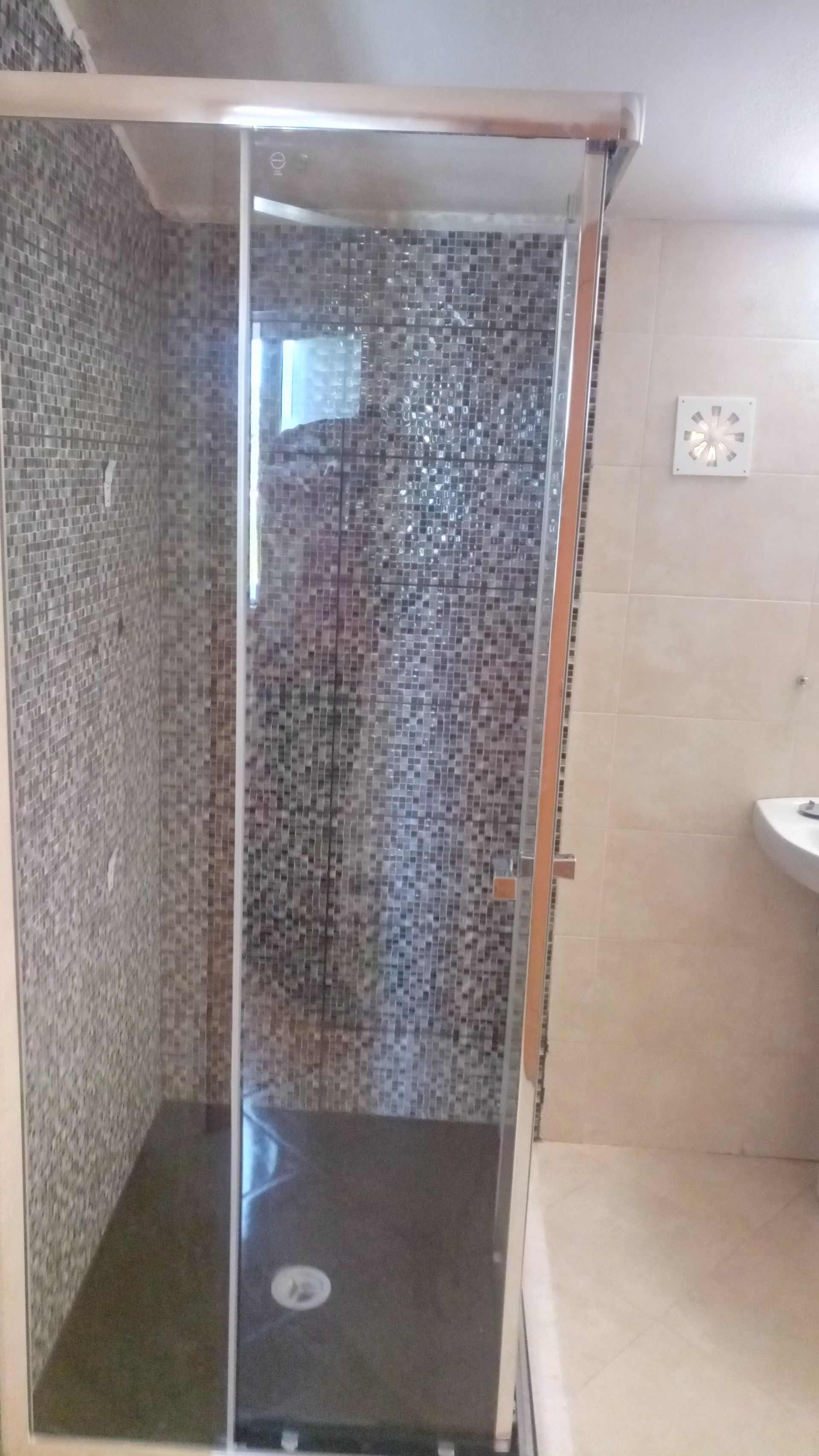 Cabine de duche com porta de vidro e paredes e chão revestidos a azulejo preto e cinzento.