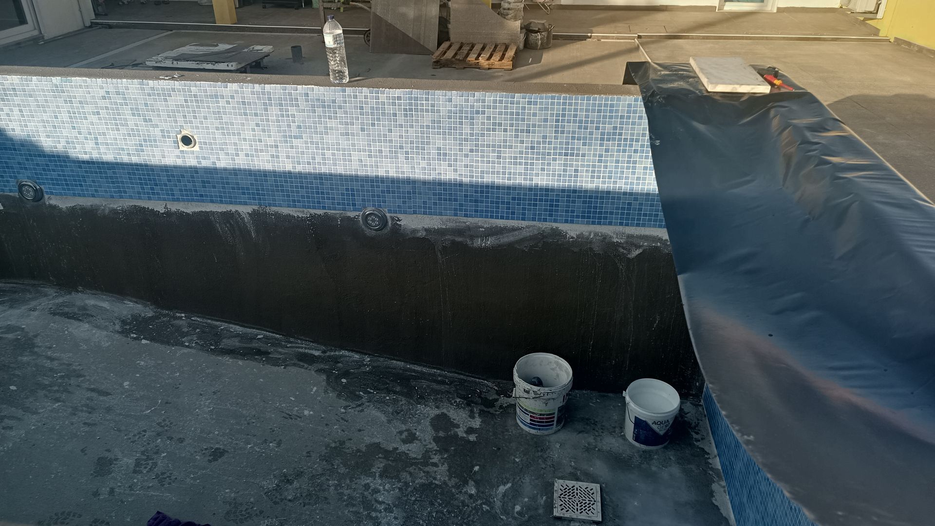 Construção de uma piscina. A parede interna da piscina possui azulejos de mosaico e está sendo revestida com uma substância escura.