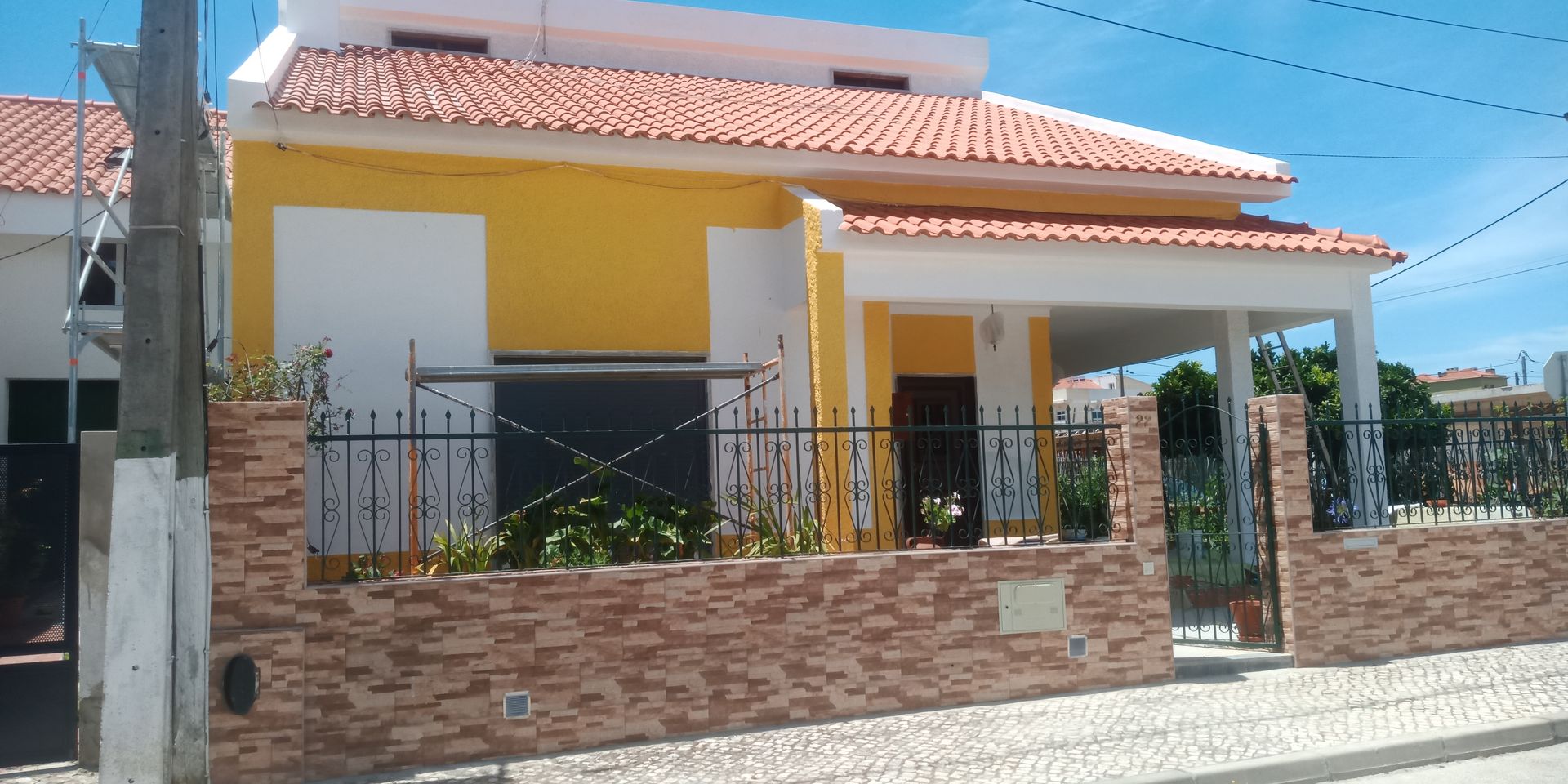 Casa amarela com telhado de telha laranja, detalhes brancos e muro de tijolos.