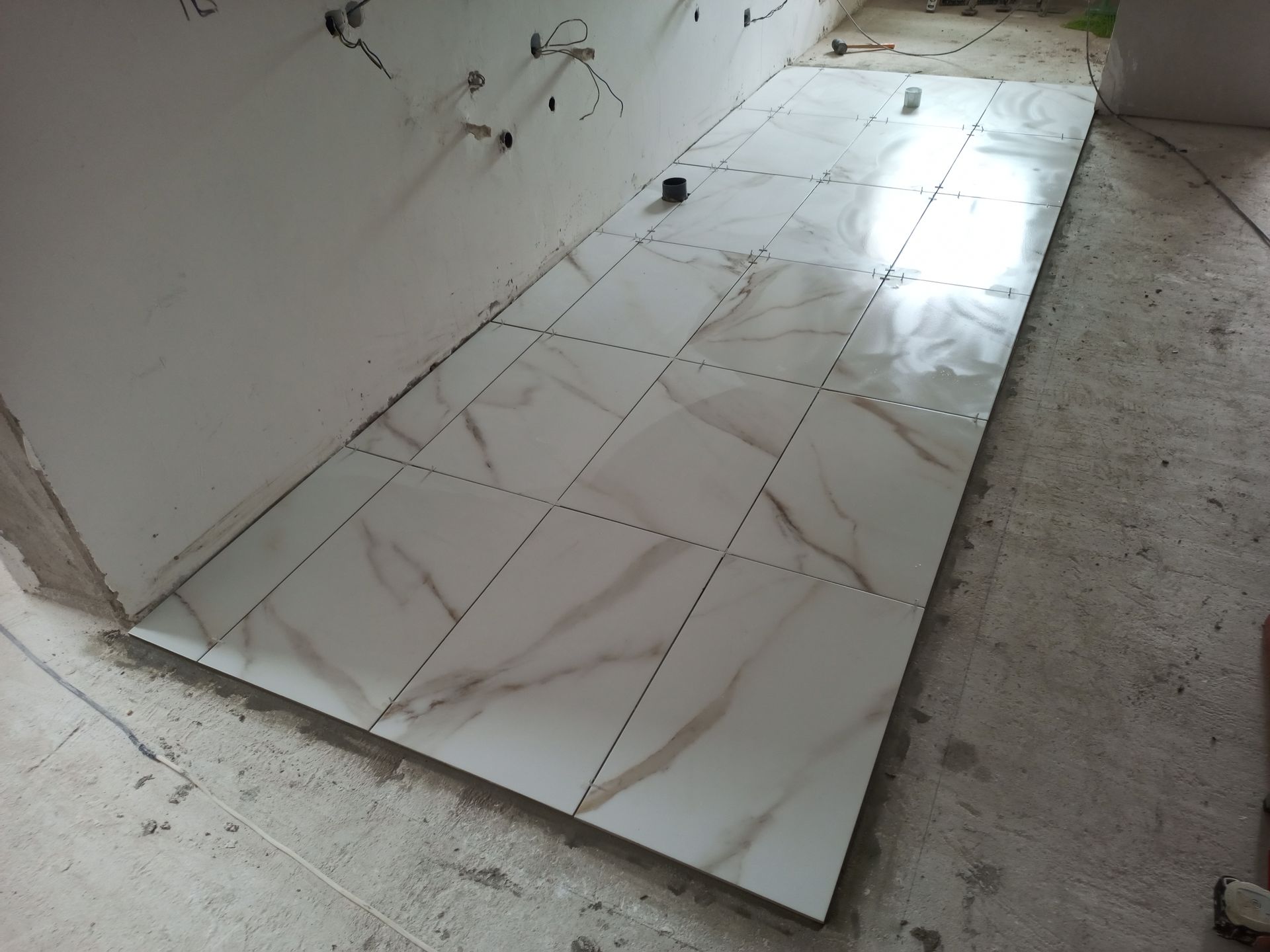 Instalação de piso cerâmico em andamento, com ladrilhos brancos imitando mármore. À esquerda, uma parede branca.