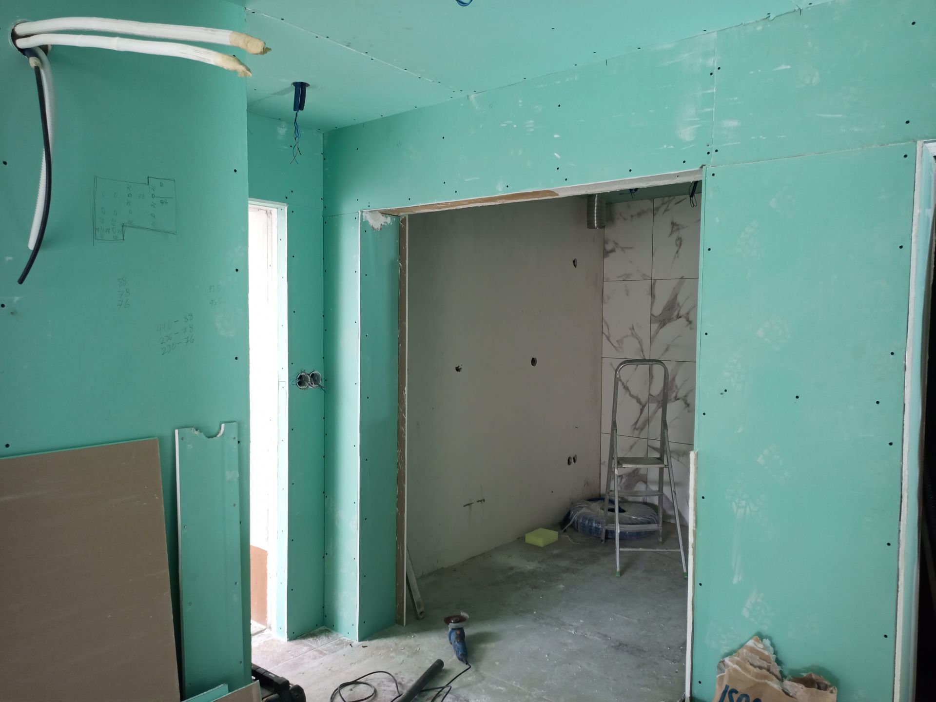Cena interna de uma obra com drywall verde, porta, escada e fios.