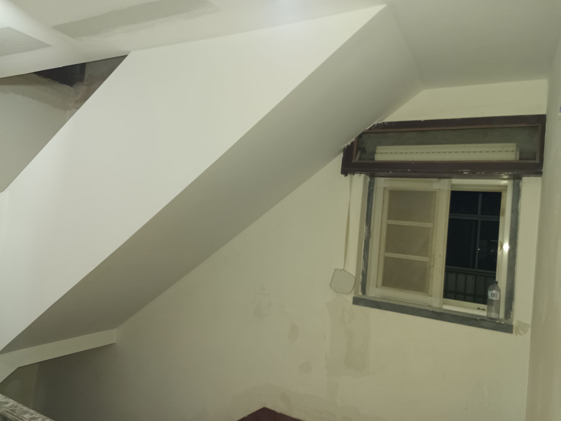 Interior com paredes brancas, janela, persiana de enrolar e seção de teto inclinada.