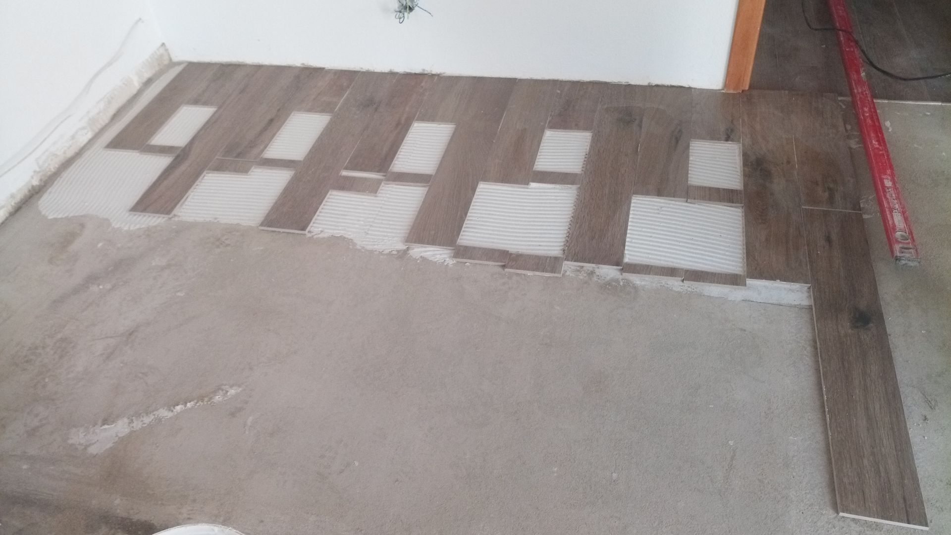 Instalação de piso em andamento: ladrilhos imitando madeira marrom assentados sobre uma base de cimento cinza; ferramentas visíveis.