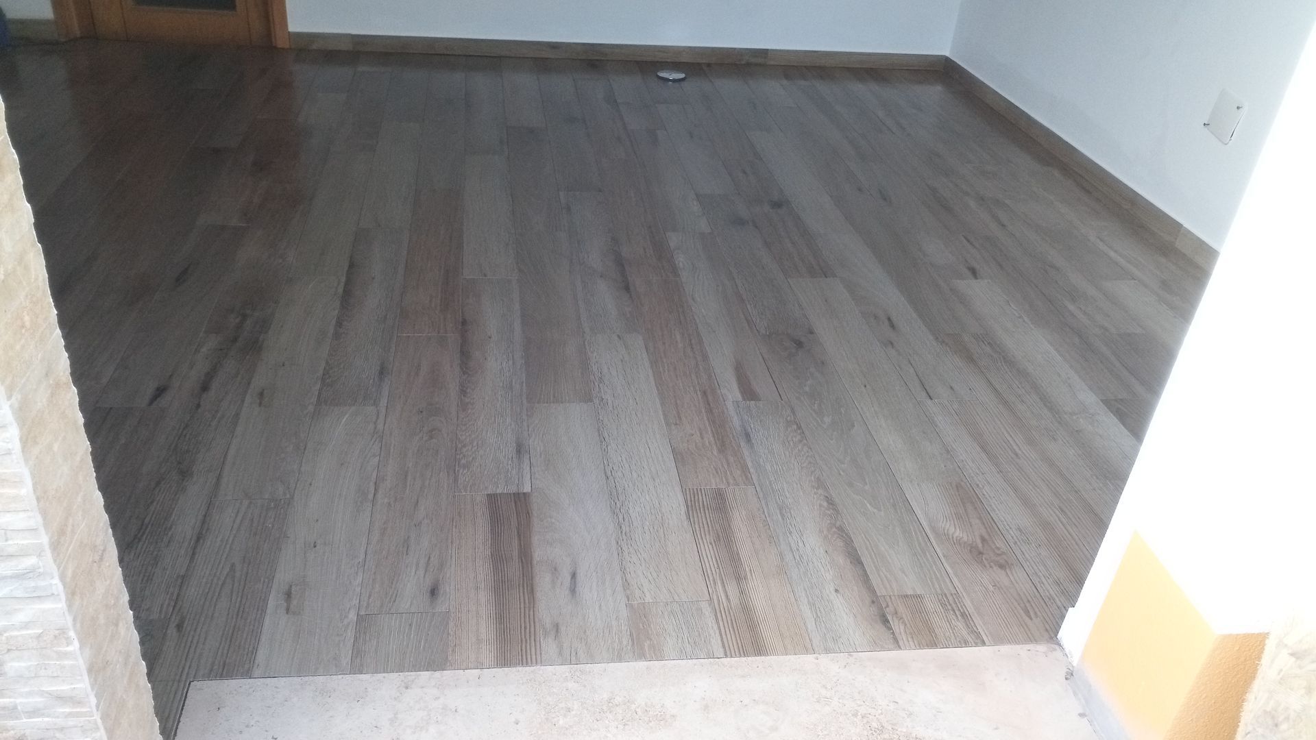 Piso de madeira maciça marrom-claro em um cômodo, delimitado por molduras escuras e paredes brancas.