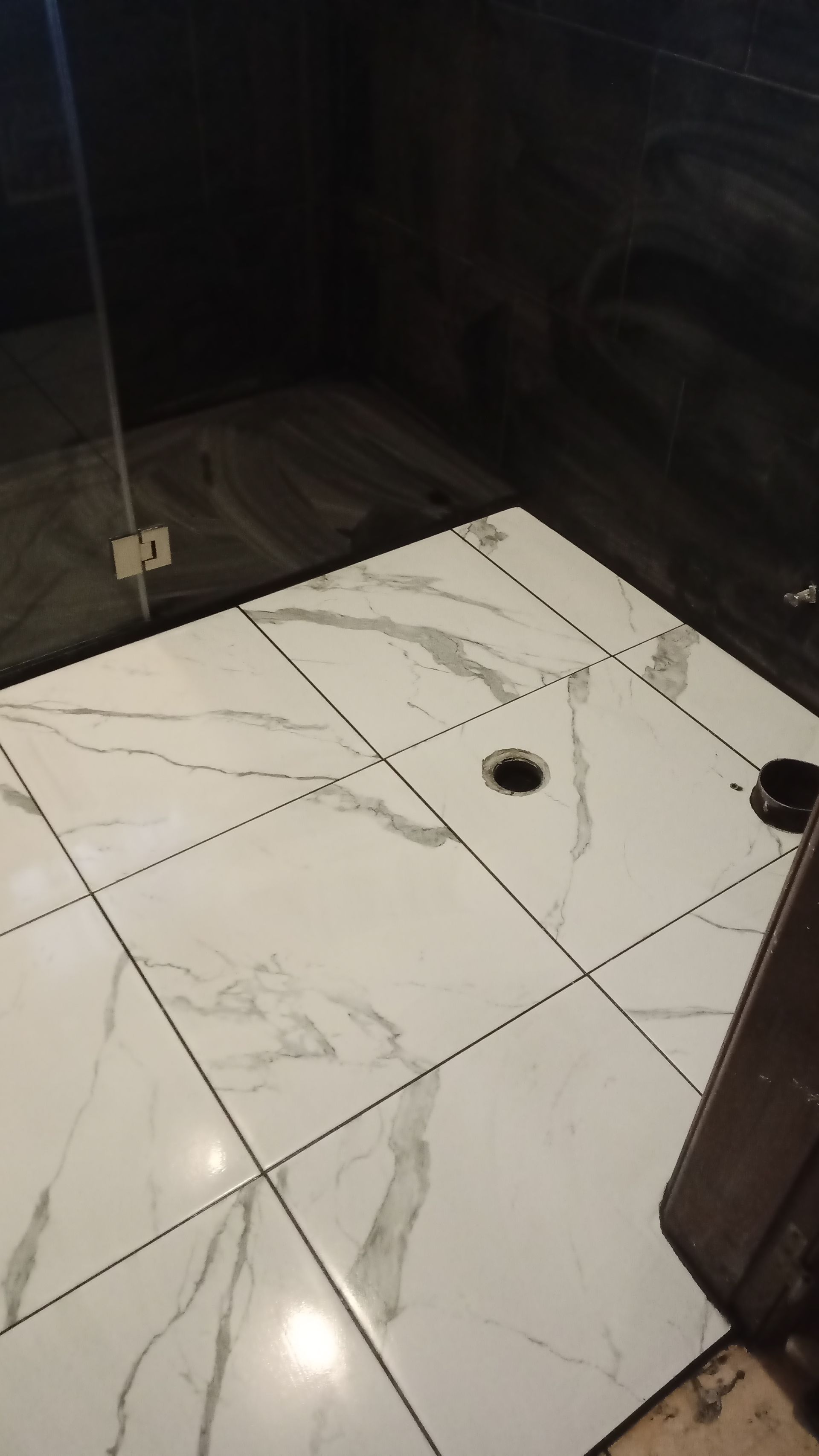 Piso do chuveiro revestido com azulejos imitando mármore branco e cinza, com paredes escuras. Ralo visível no centro.
