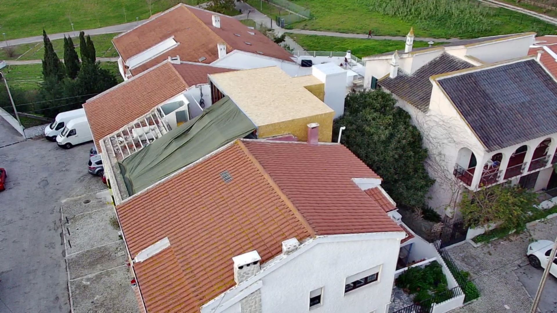 Vista aérea de casas com telhados de telha vermelha, paredes brancas e alguns veículos estacionados na rua.