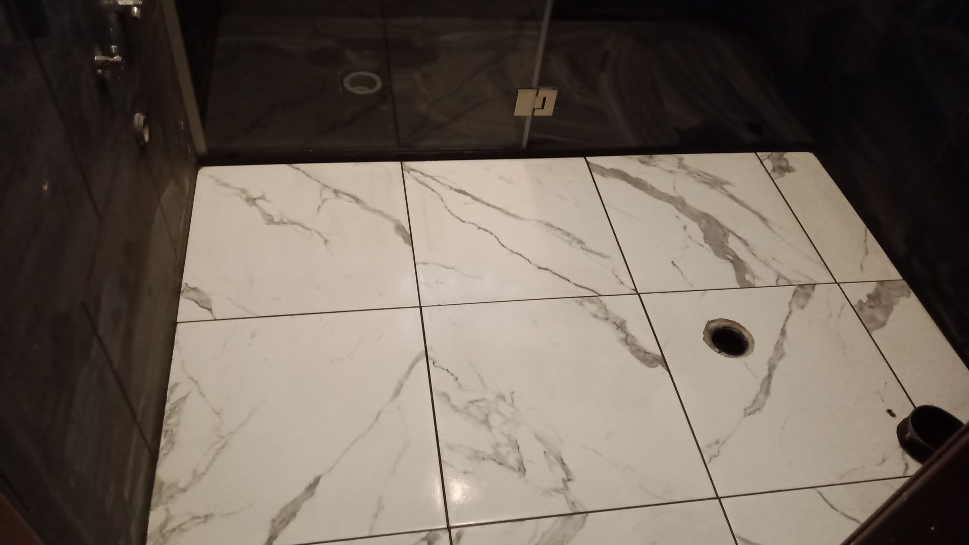 Piso do chuveiro revestido com azulejos brancos com veios cinza, ralo central. Azulejos de parede em tons de cinza escuro e preto.