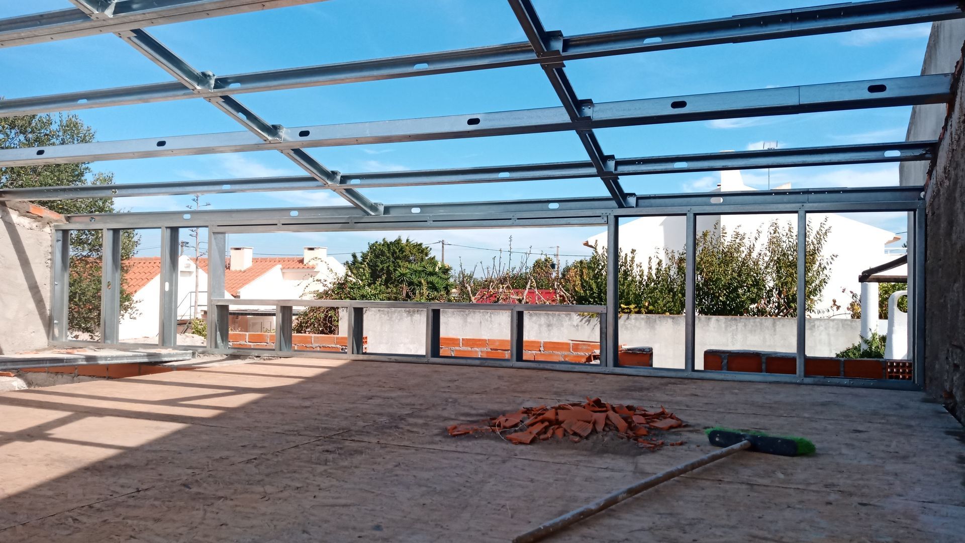 Estrutura metálica de um edifício em construção, com laterais abertas, piso de concreto e vista para o céu e casas.
