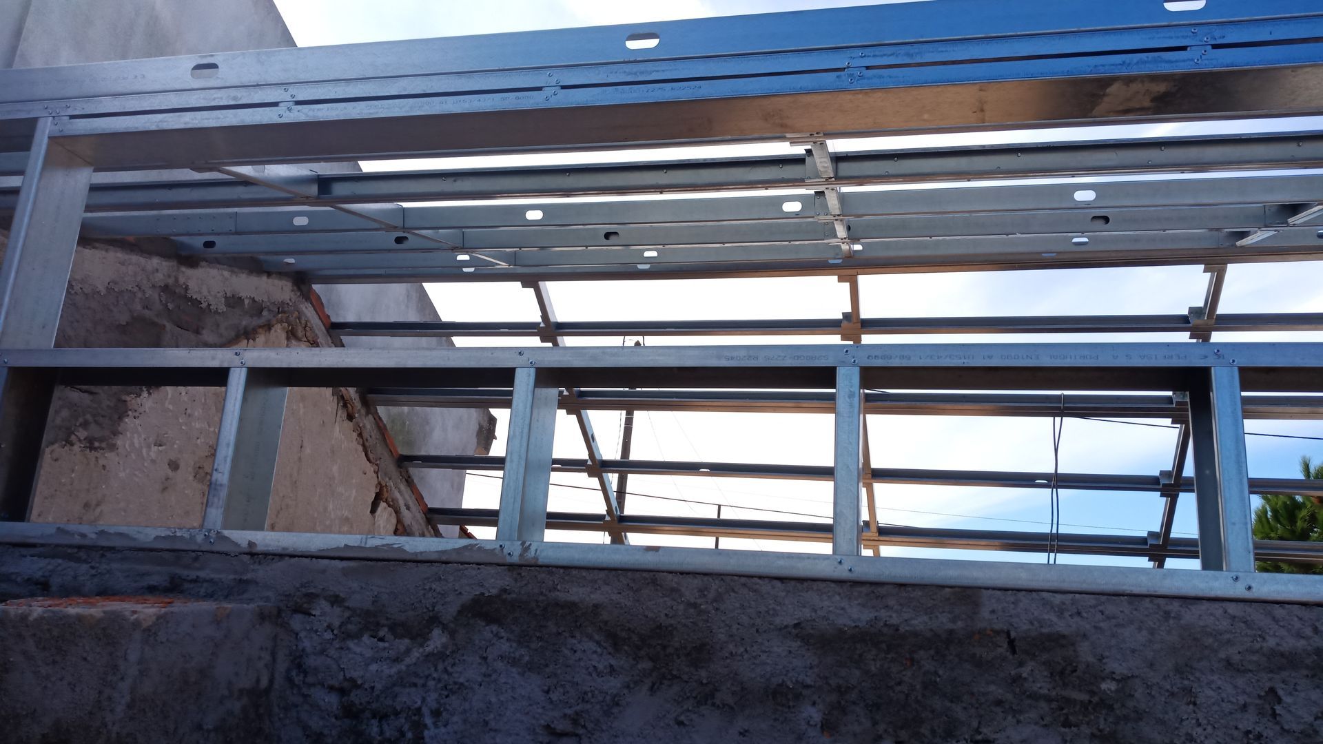 Estrutura metálica para telhado ou forro; fixada a uma parede de concreto; permite a entrada de luz natural através da estrutura.