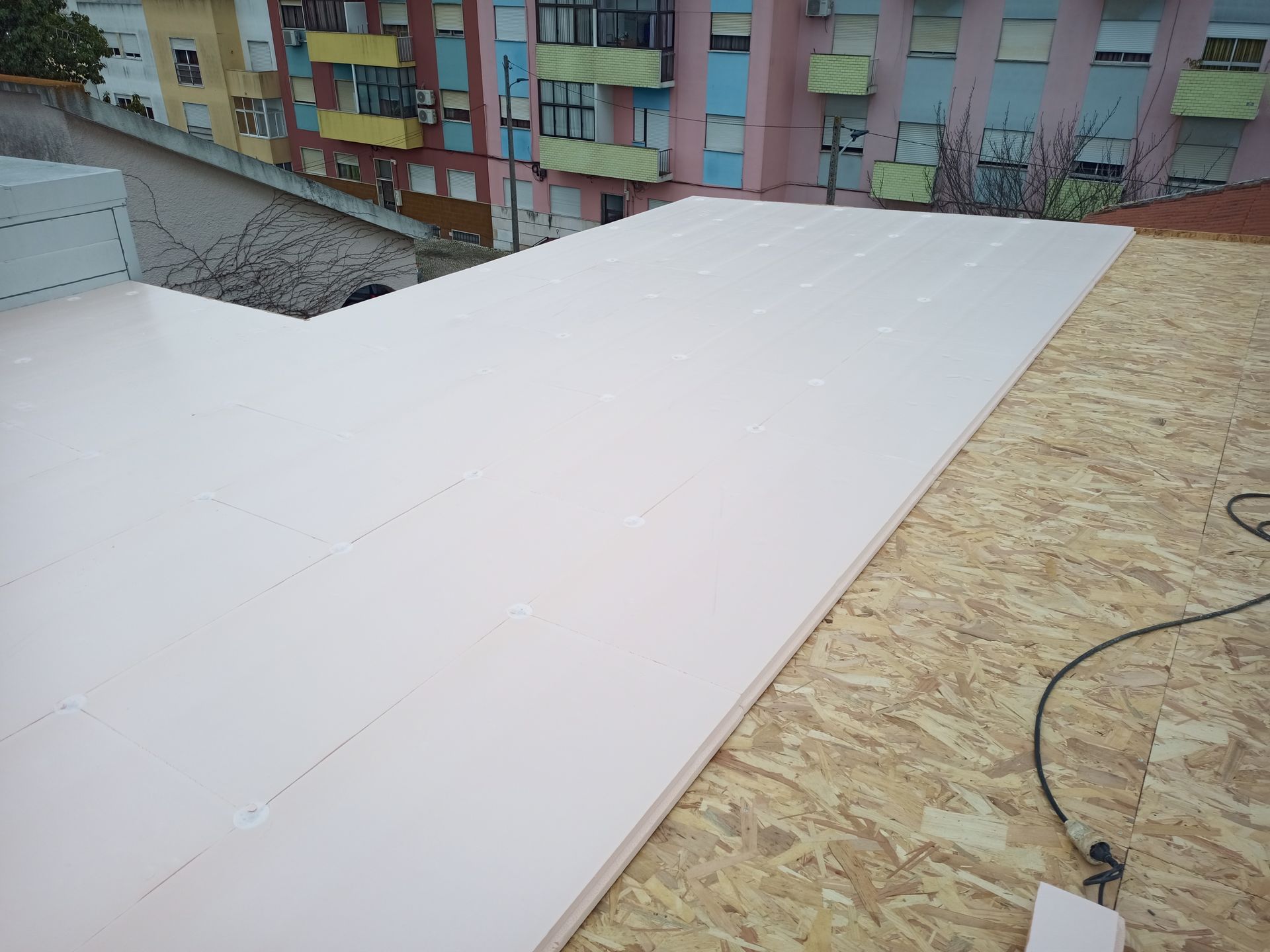 Painéis isolantes rosa sendo instalados em um telhado de OSB, com prédios de apartamentos ao fundo.